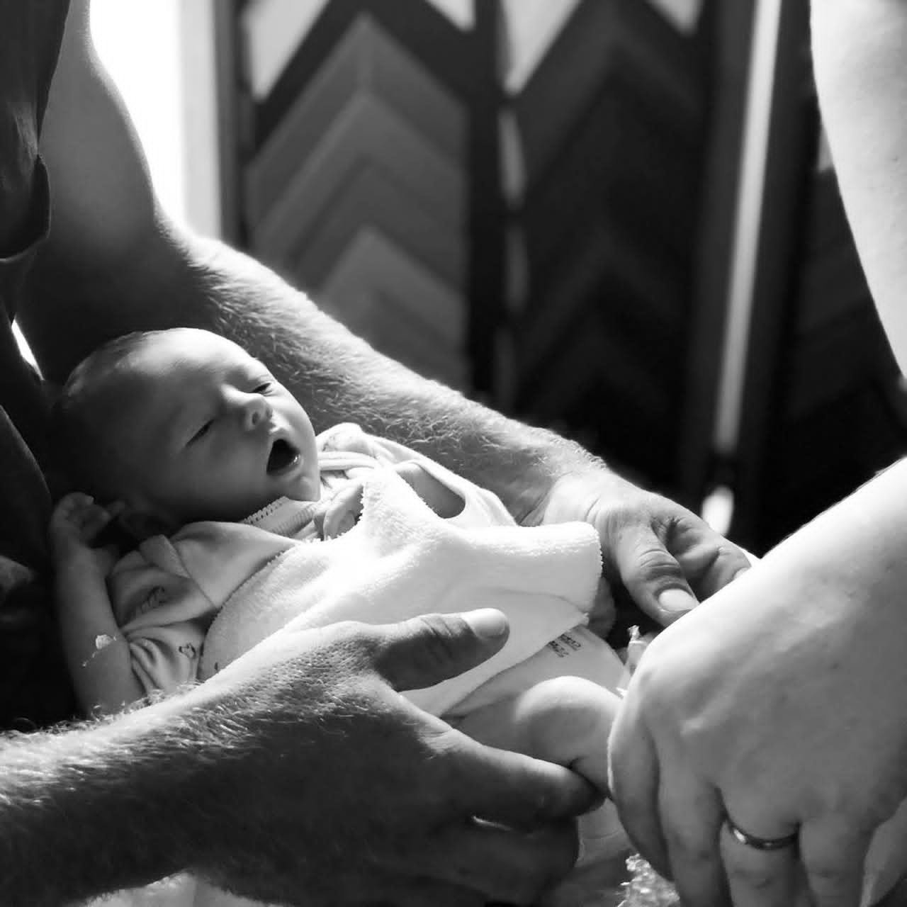 Black and white photo of a newborn baby being held and supported by multiple adult hands, with a patterned background.
