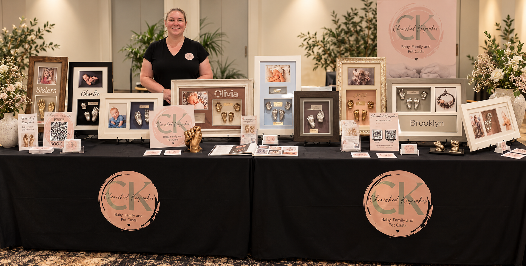 A woman standing behind a display table at a craft fair showcasing baby, family, and pet casts in various display frames and sculptures. The table is decorated with flowers and promotional signs.