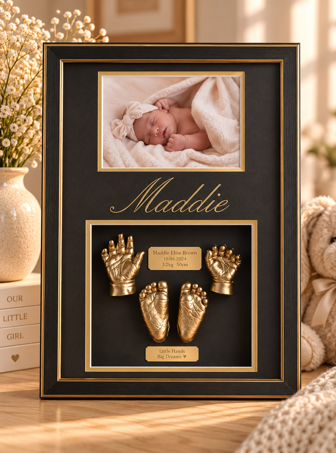 A framed baby keepsake with a photo of a sleeping newborn girl at the top, gold baby hand and foot casts with plaques containing name, birth date, weight, and height, and decorative elements including the name "Maddie" in elegant script.