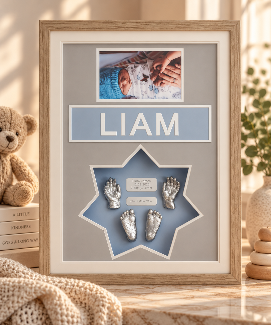 A shadow box frame with a baby photo at the top, the name Liam in large letters, and silver baby handprints and footprints inside a star-shaped cutout, along with birth details and a birth quote.