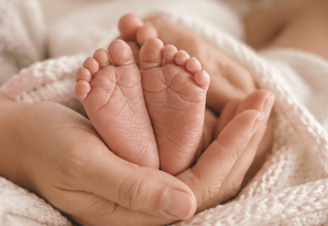 Close-up of an adult hand gently holding a newborn baby's tiny feet, wrapped in a soft, cream-colored blanket.