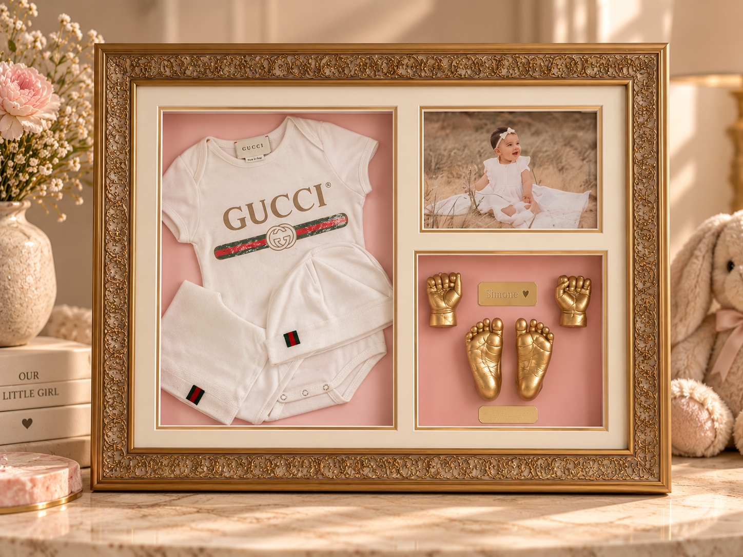 Decorative photo frame with a baby outfit, a photograph of a baby girl, and golden baby feet and hands keepsakes, surrounded by flowers and plush toys.