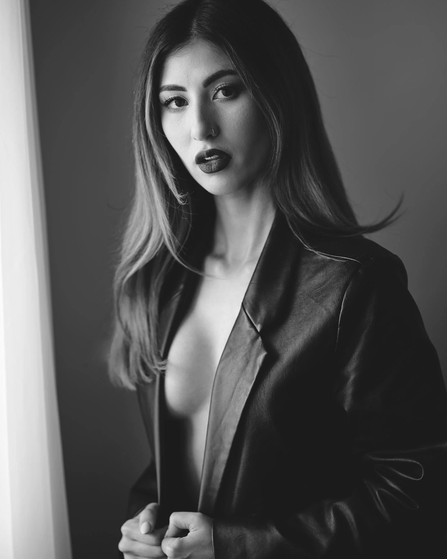 Black and white portrait of a woman with dark lipstick and a nose ring, wearing a leather jacket, looking into the camera in front of a plain wall.