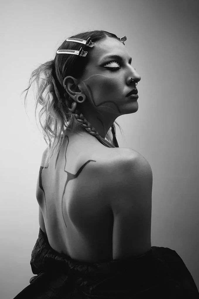 Black and white photo of a woman with tattoos on her back in profile view, with hair held back by hair clips, wearing makeup and piercings.