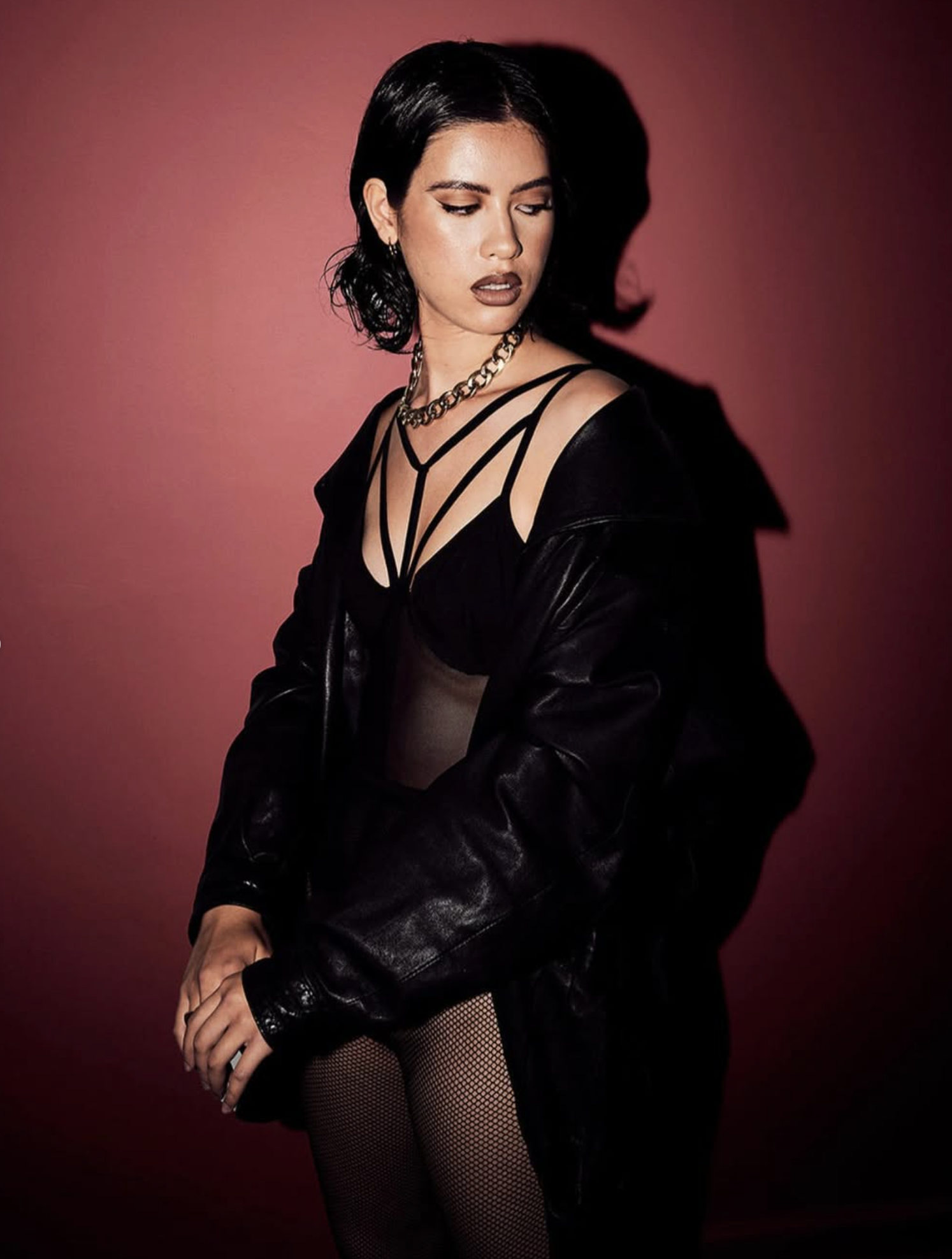 A woman with short black hair wears a black strappy top, a black leather jacket, fishnet stockings, and chunky chain jewelry, standing against a red background.