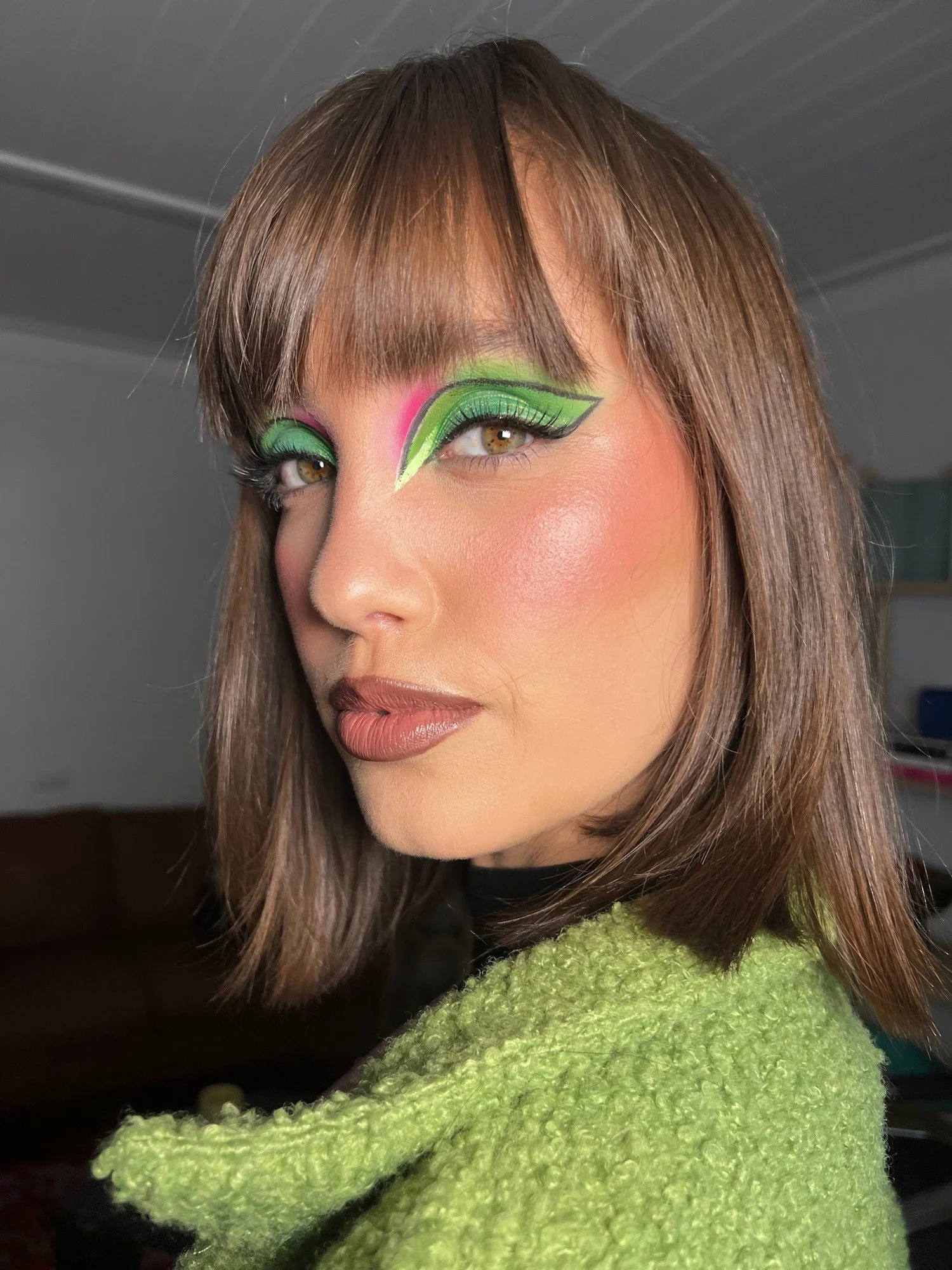 Close-up of a woman with colorful green and pink eye makeup and brown hair, wearing a bright green fuzzy sweater.