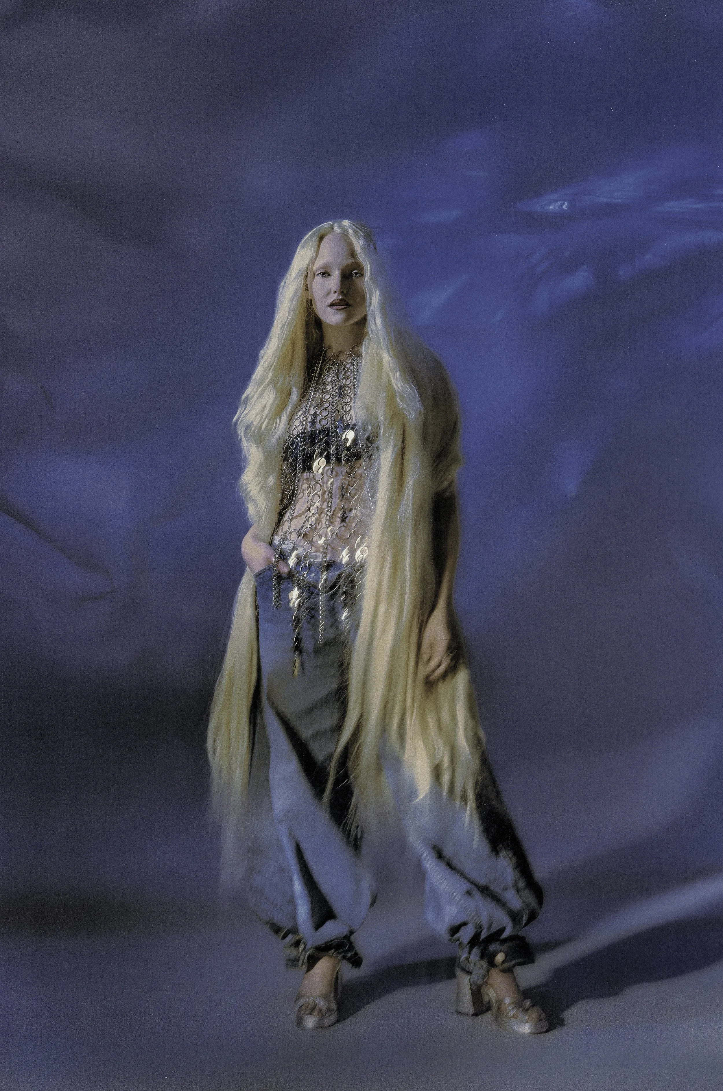 A woman with long blonde hair in a fashion pose, wearing a light-colored, layered outfit with chains and accessories, and high-heeled sandals, against a dark background with blueish lighting.