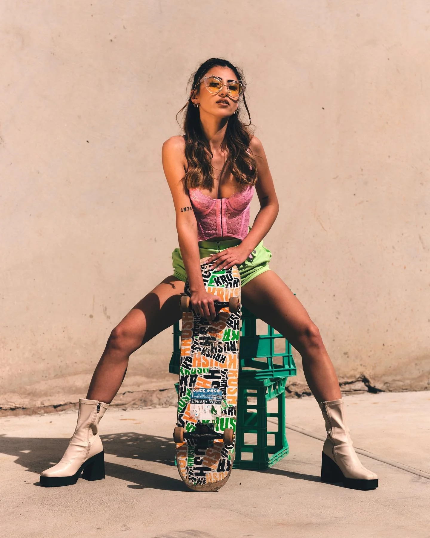 A woman with long dark hair and sunglasses sitting on green crates with her legs apart. She is holding a skateboard with colorful text, wearing a pink corset, neon green shorts, and white high-heeled boots, outdoors against a plain beige wall.