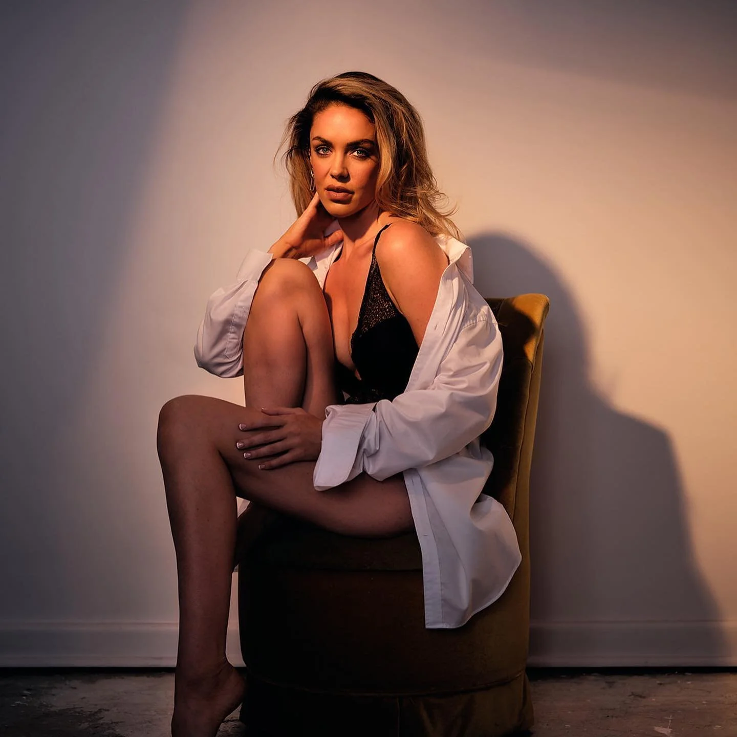 A woman with blonde hair in a black lace lingerie, white shirt draped over shoulders, sitting on a brown chair against a plain wall with soft lighting.