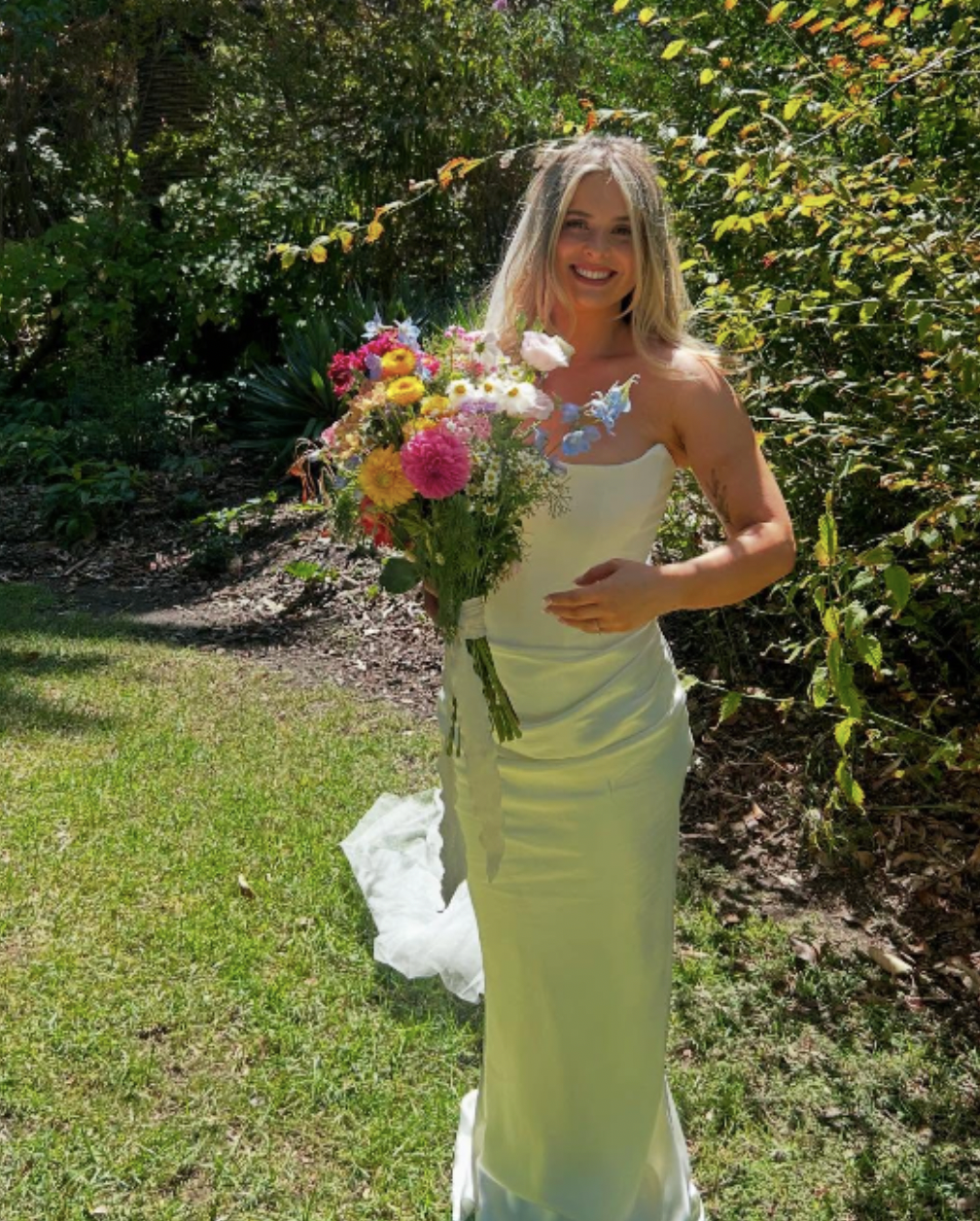 A woman in a white wedding gown holding a colorful bouquet of flowers in an outdoor garden setting, smiling at the camera.