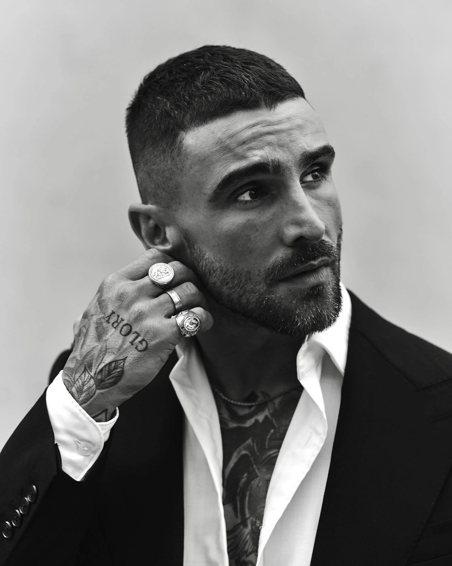 A black and white portrait of a man with short dark hair and a beard, wearing a suit jacket and a white shirt. He has several rings on his fingers, a tattoo on his hand, and is looking to the side while adjusting an earring or earring area.