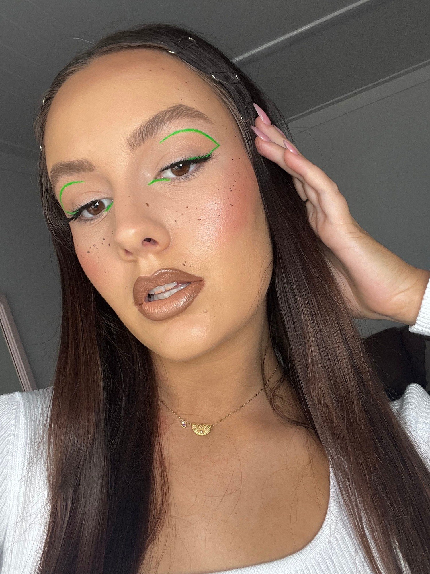 A woman with long brown hair, wearing a white top and gold jewelry, is touching her face with her hand. Green eyeliner is drawn on her eyelids.