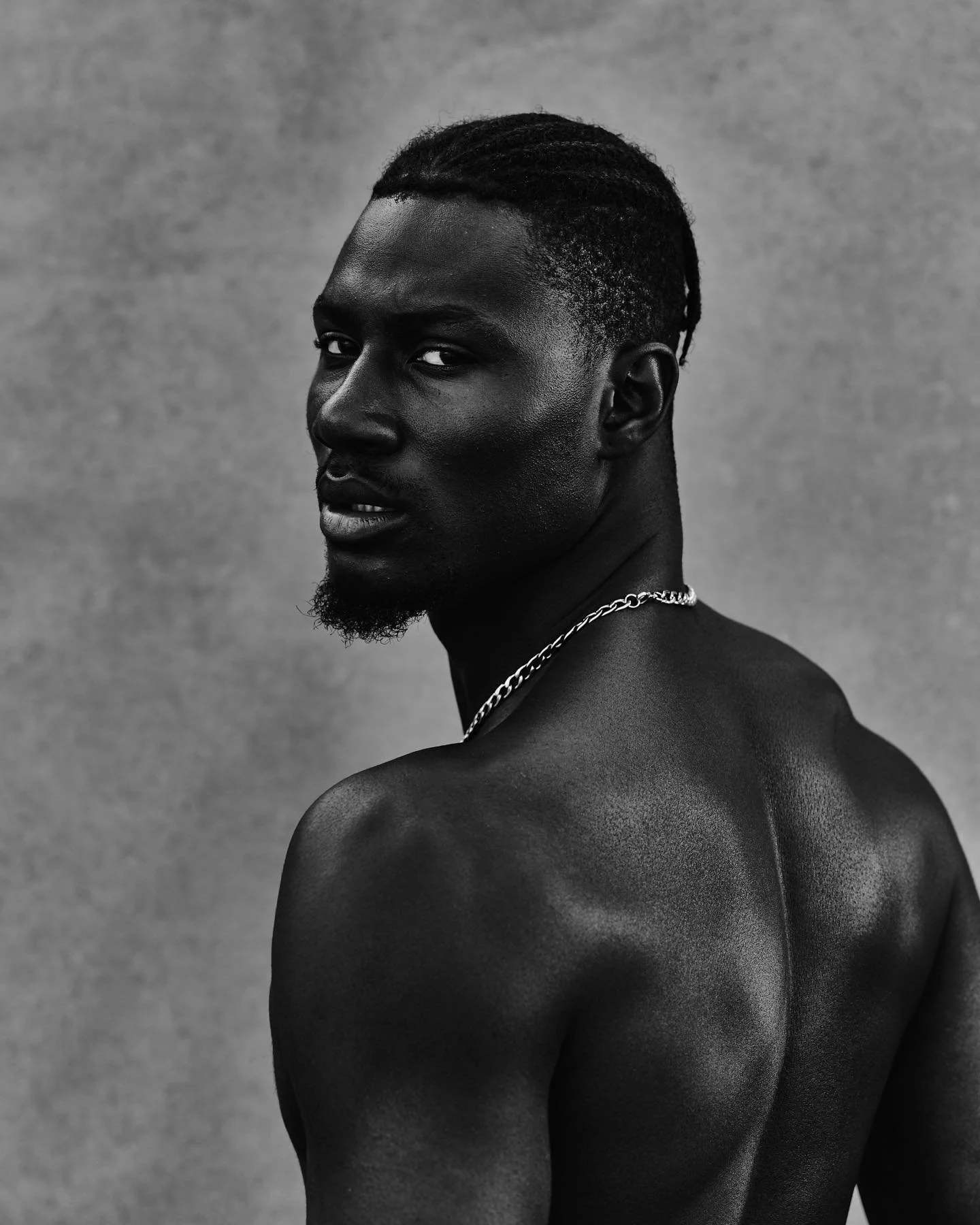 Black and white portrait of a shirtless young man with short dreadlocks, a goatee, and a chain necklace, looking over his shoulder against a plain textured background.