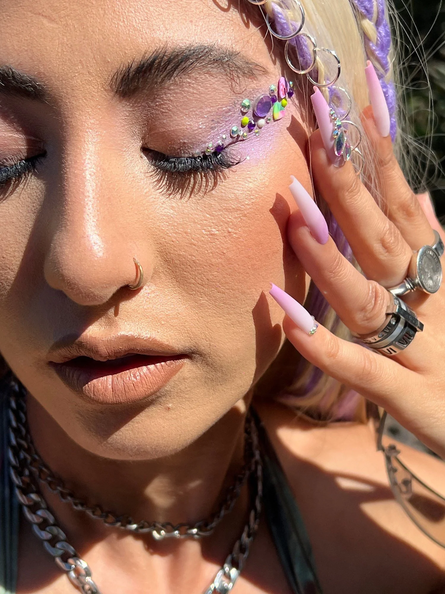 Close-up of a woman's face with makeup, jewelry, and long pink nails. She has her eyes closed and is touching her face with her hand.