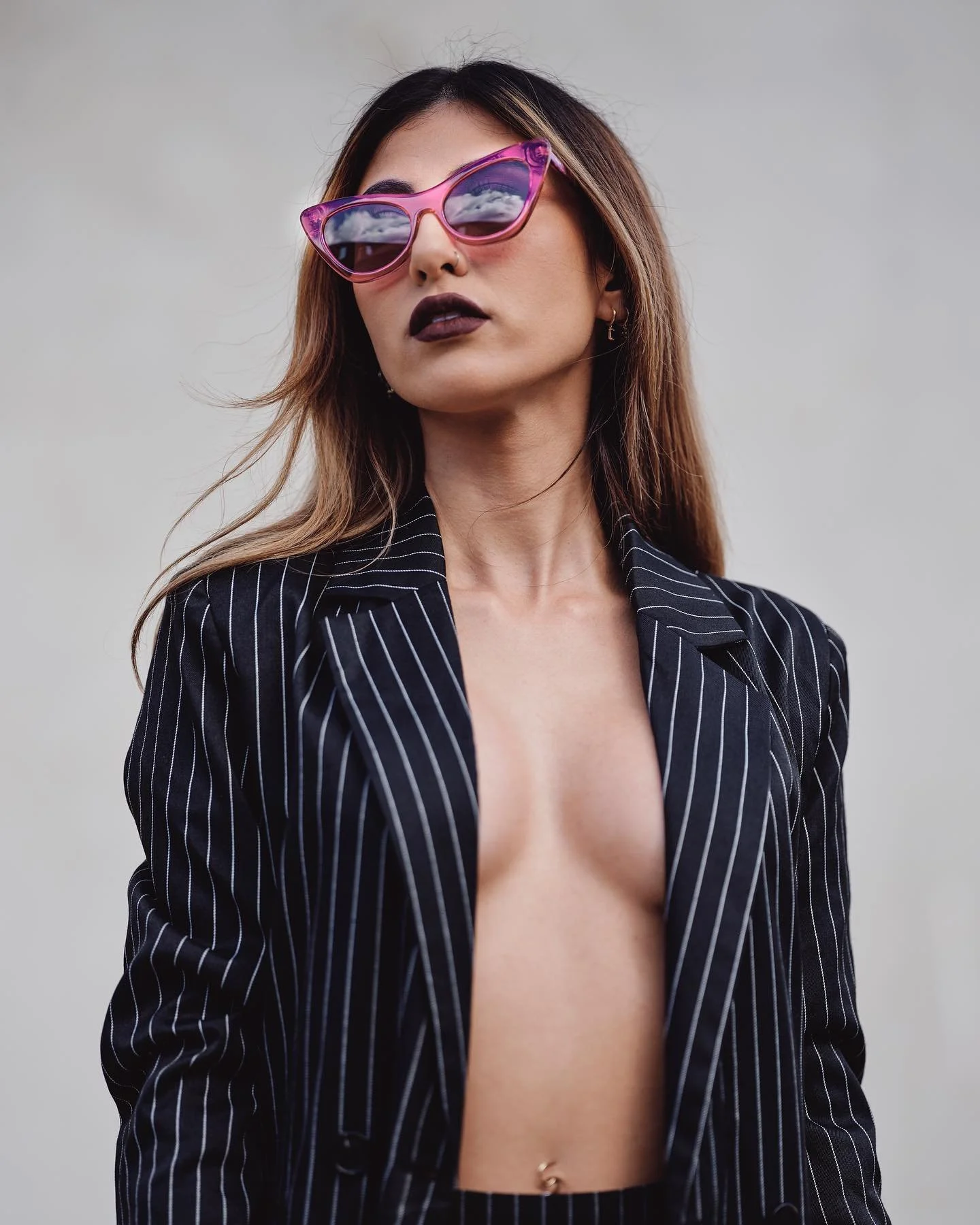 Young woman with long brown hair wearing pink sunglasses, dark lipstick, and a black pinstripe blazer with no shirt underneath, standing against a plain gray background.