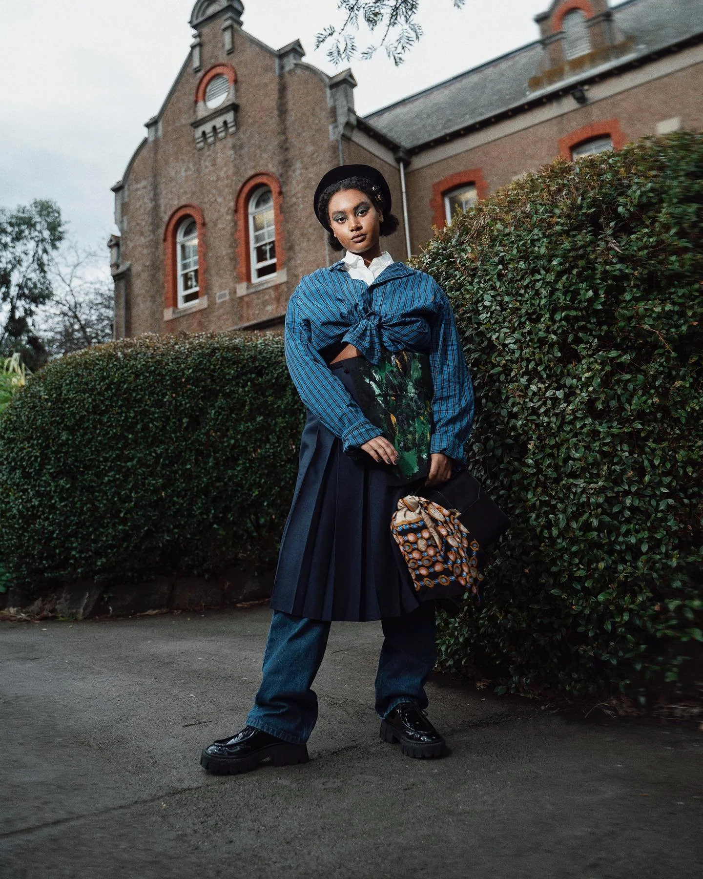A woman stands outdoors in front of a hedge and a brick building, wearing a black hat, white shirt, and blue checkered jacket tied at the waist, with bag and jeans.