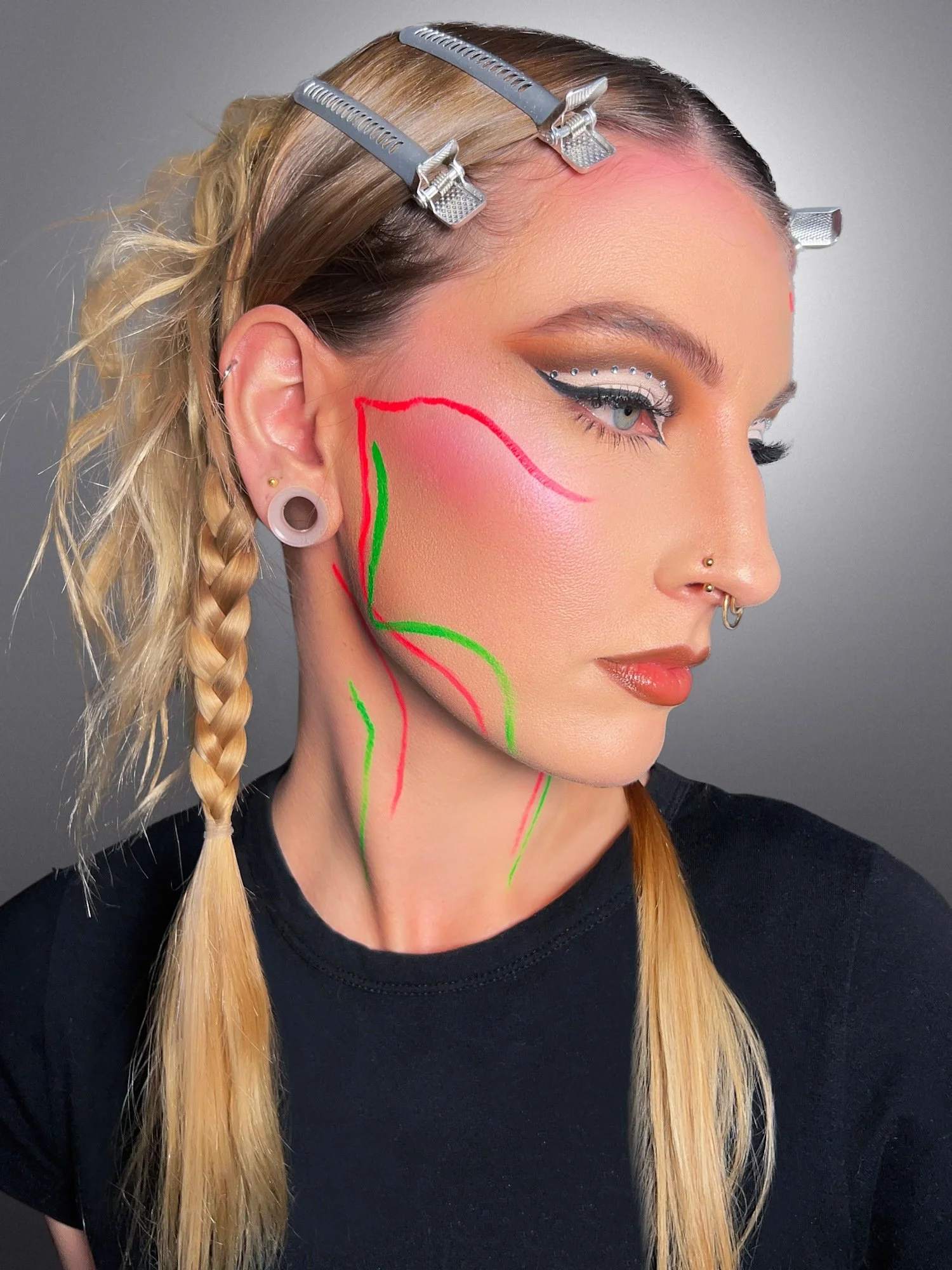 A woman with blond hair styled in braids, wearing makeup including eyeliner and rhinestones, has surgical clips on her hair, and colorful markings on her face and neck.