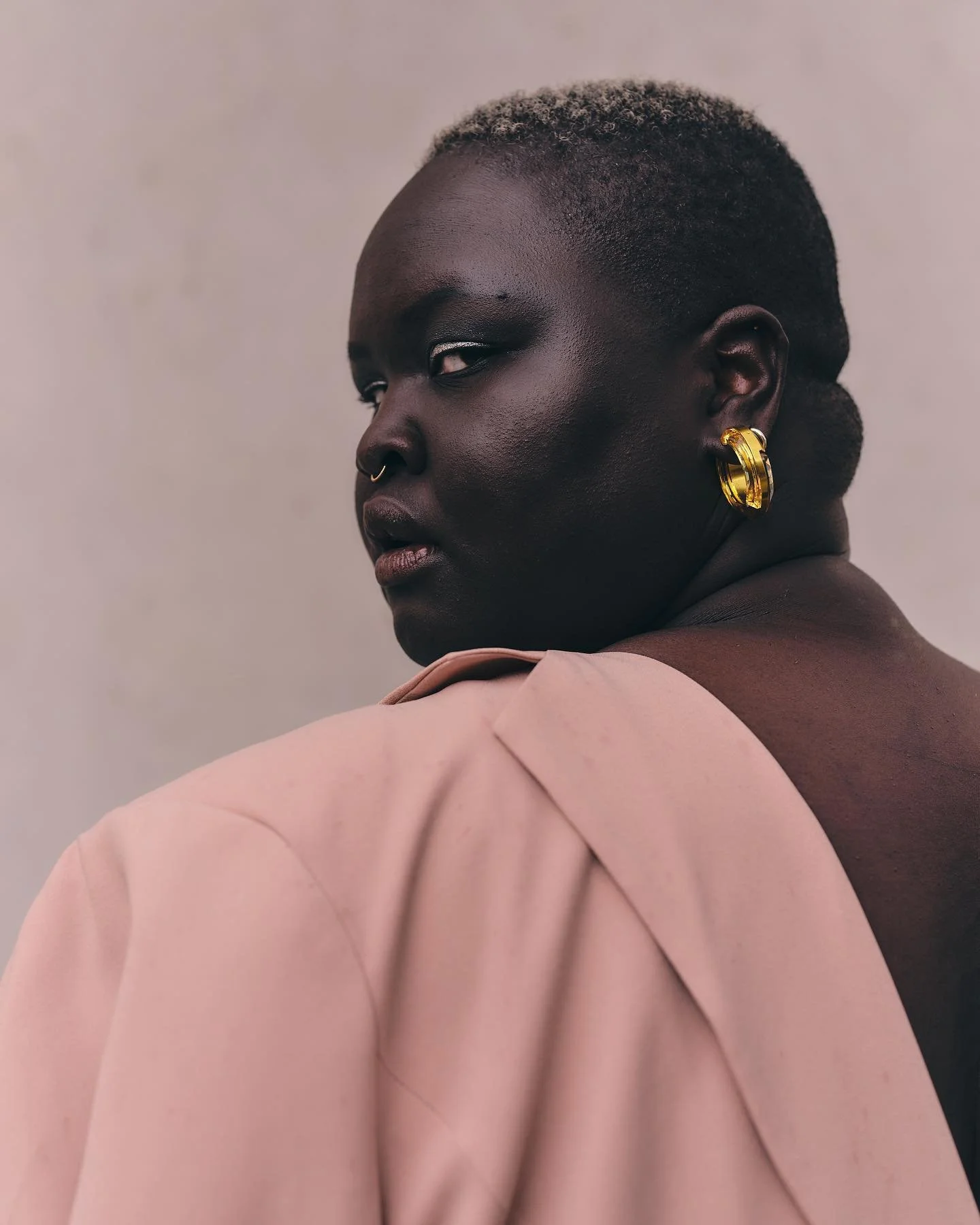 A profile portrait of a Black woman with short, curly hair and gold earrings, wearing a pink blazer, against a neutral background.