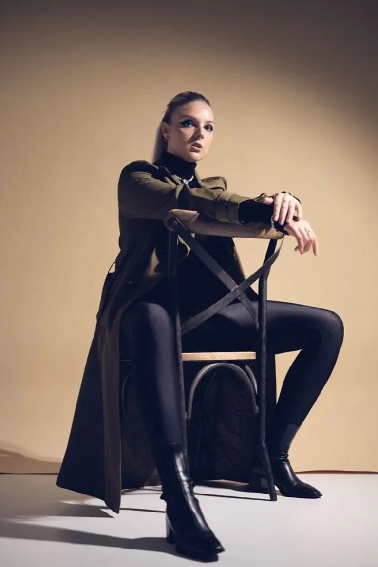 A woman in black leggings, a black turtleneck, and a brown trench coat sitting on a black chair against a plain beige background.
