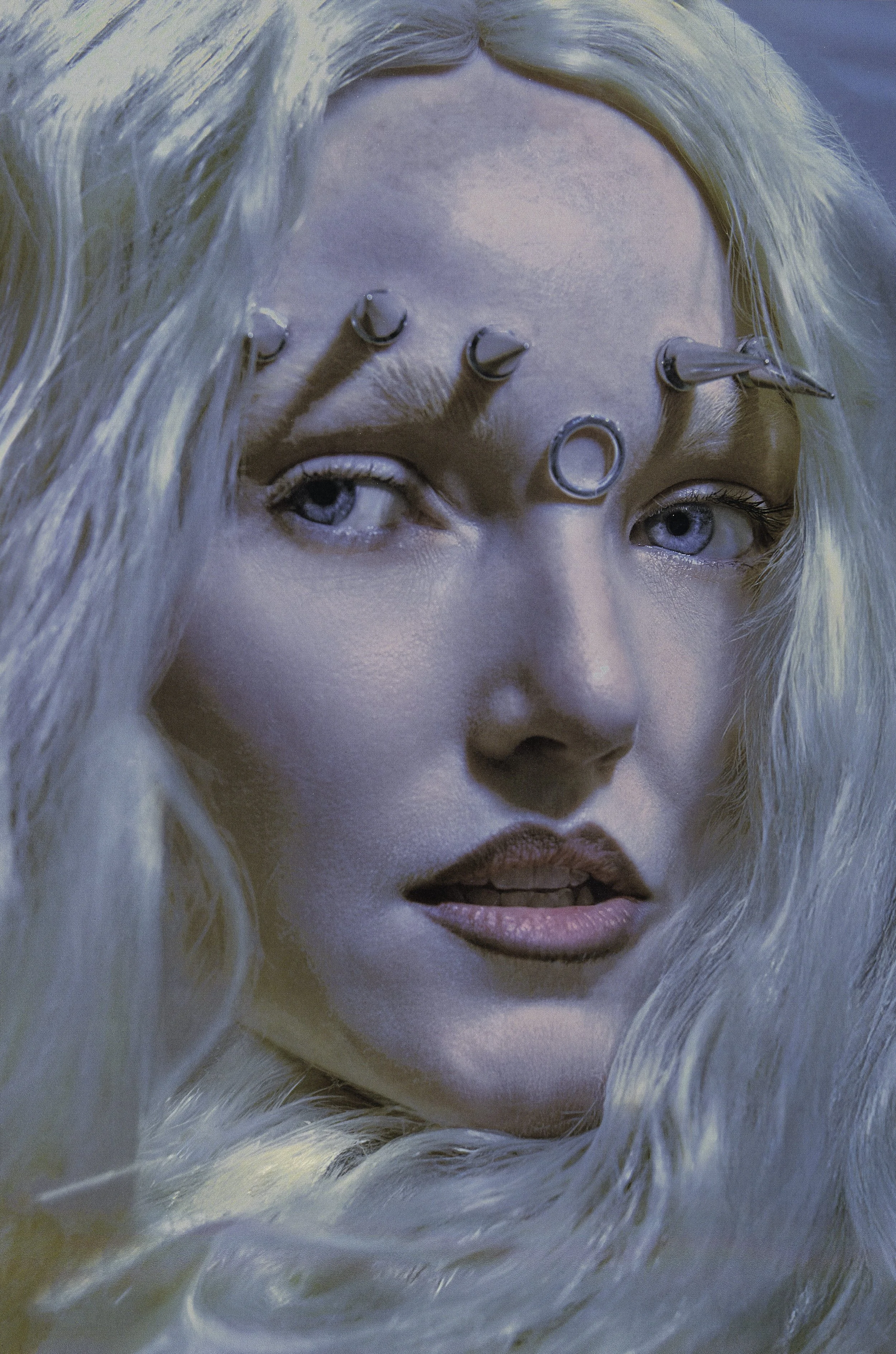 A close-up of a female model's face with long, wavy platinum blonde hair, with multiple small metallic nails embedded in her forehead, and her lips slightly parted.
