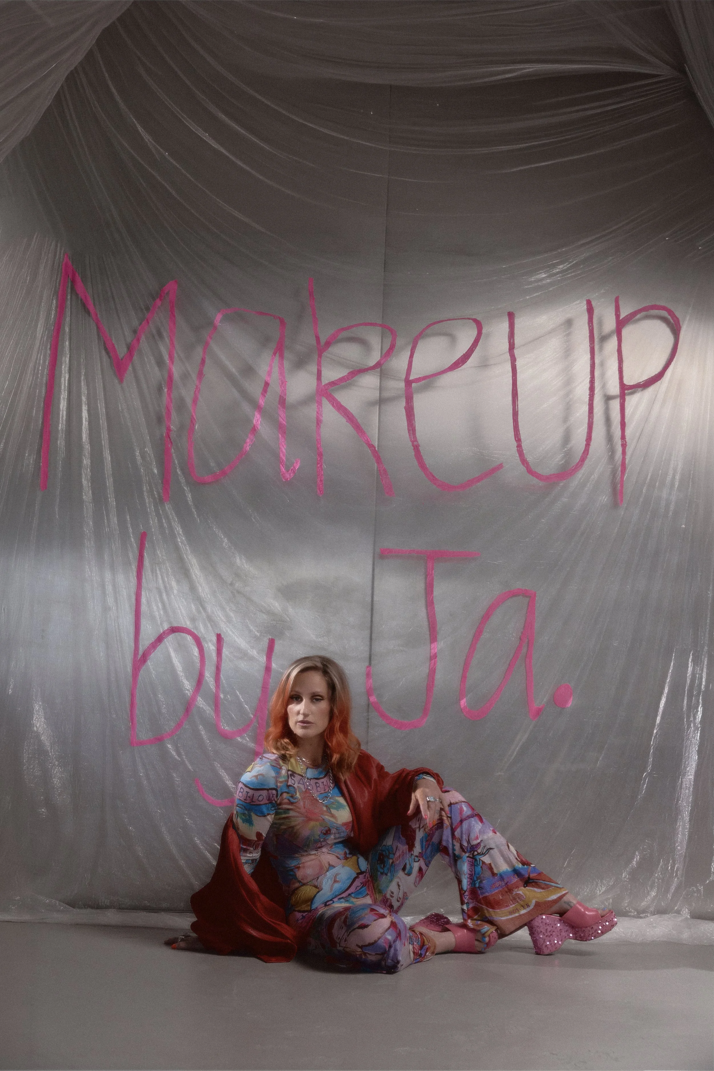 A woman with curly orange hair sitting on the floor in front of a shiny silver backdrop with large pink handwritten text. She is wearing colorful, patterned pajamas, a red velvet jacket, and pink platform shoes.