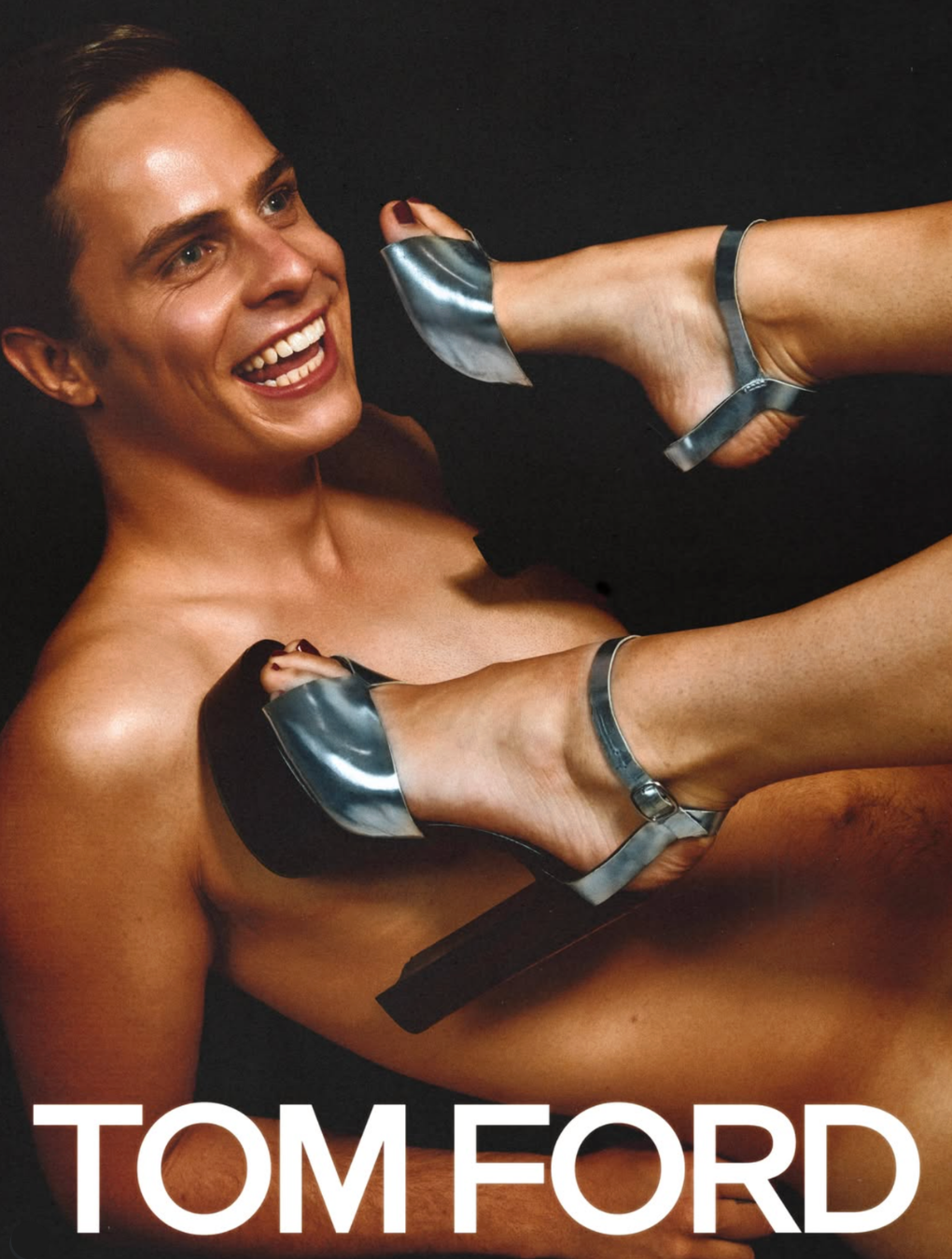 A woman with short hair and a big smile, lying down. She is nude, with metallic high-heeled shoes on her feet. The background is black, and the text 'TOM FORD' is at the bottom of the image.