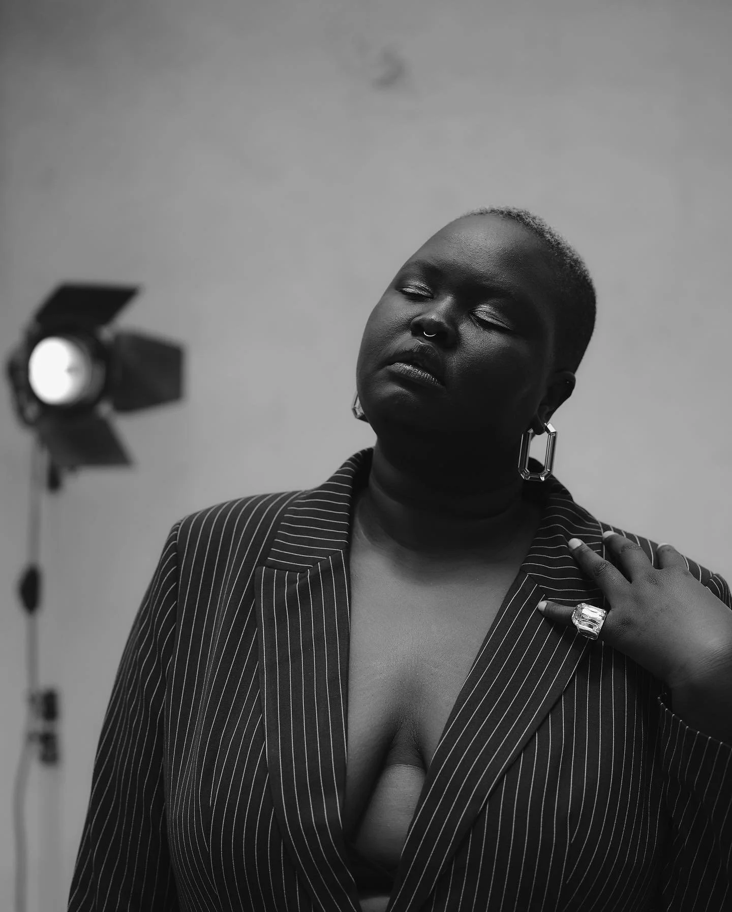 Black and white photo of a woman with closed eyes, short hair with a light-colored edge, wearing large earrings, a pinstriped blazer, a ring, a watch, and a nose ring, with a photo light in the background.