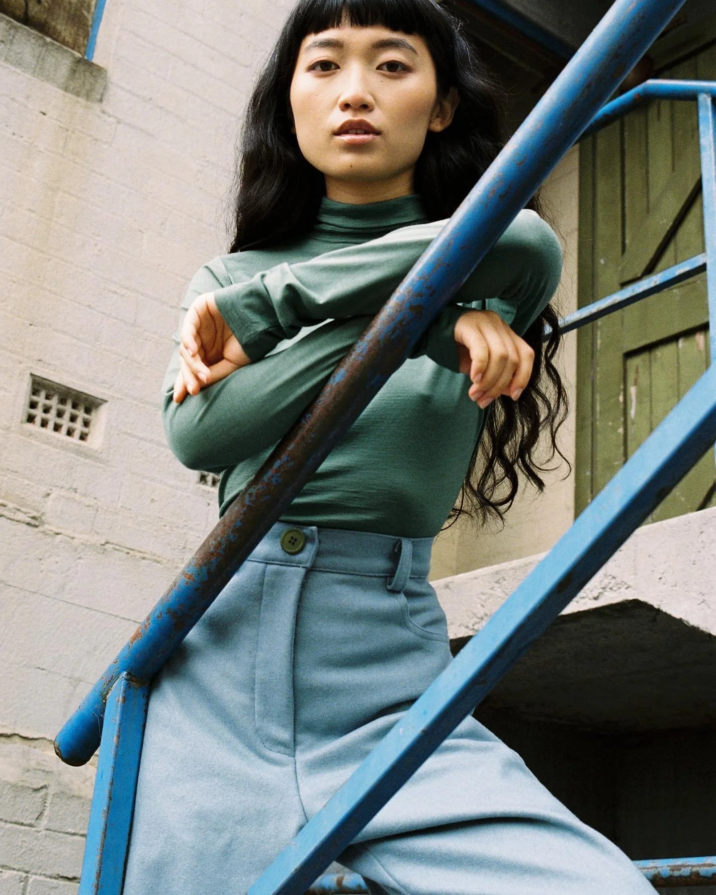 A young woman with long dark hair and bangs, wearing a green long-sleeve top and high-waisted light blue pants, leaning on a blue metal staircase railing inside a building with brick walls and green door.