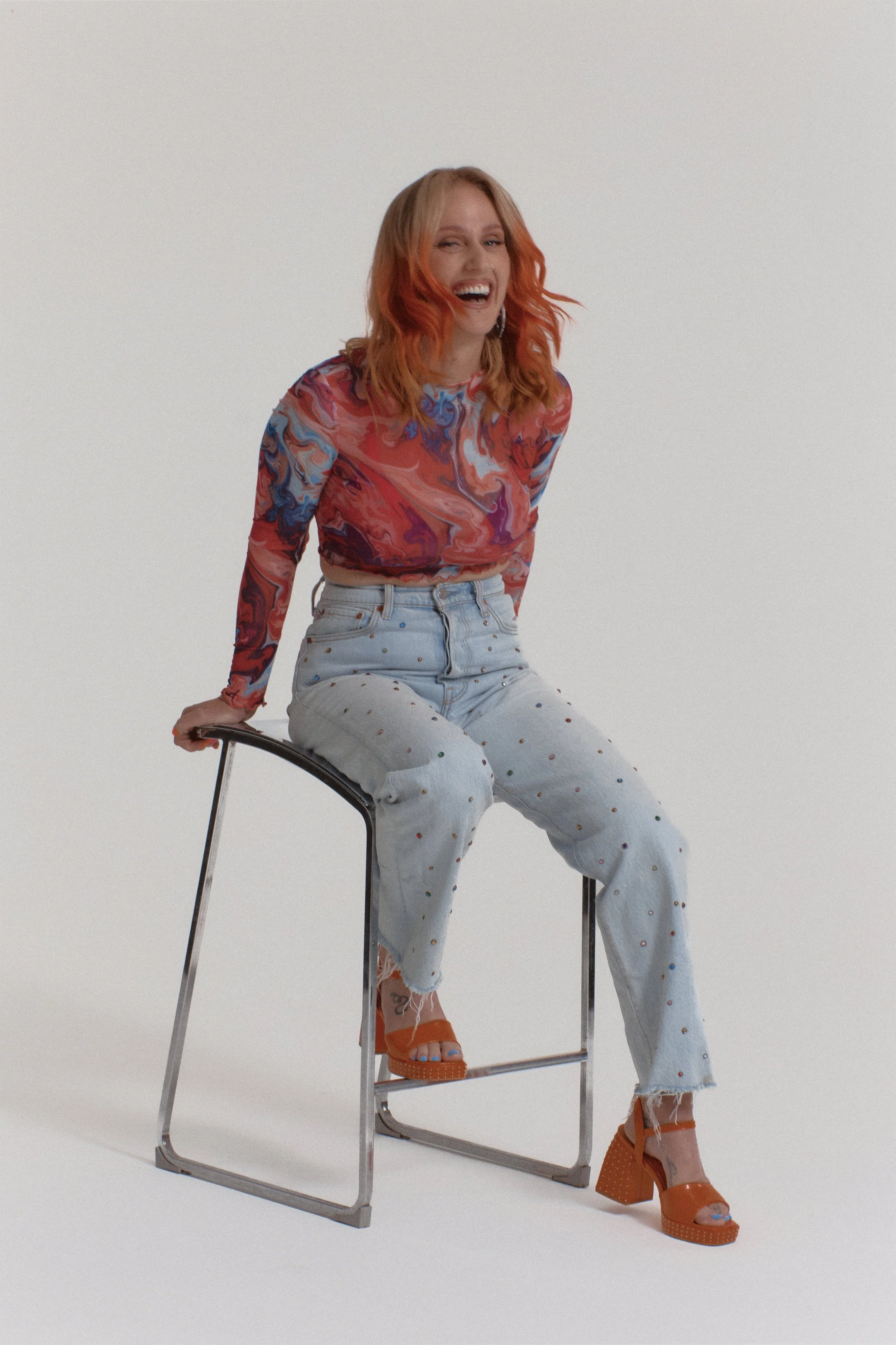 A woman with orange hair sitting on a black metal chair, laughing. She is wearing a colorful, swirling patterned long-sleeve top, distressed high-waisted jeans with small painted dots, and orange chunky high-heeled sandals. The background is plain white.