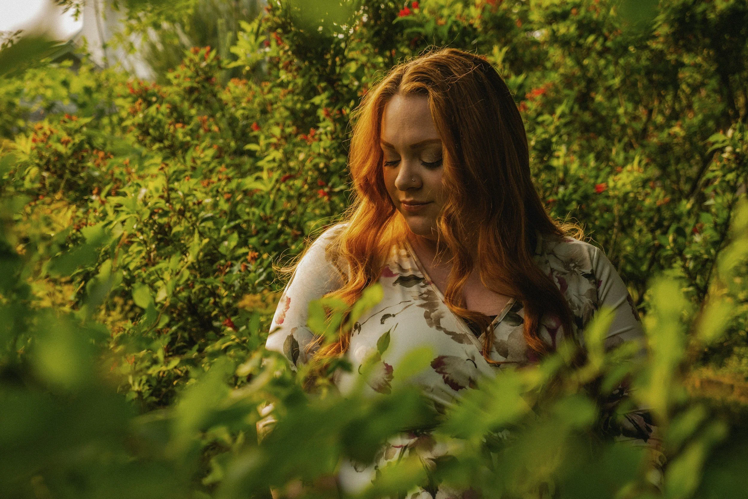 A woman with long red hair and a nose piercing stands among green bushes, looking downward with her eyes closed, during a sunny day.