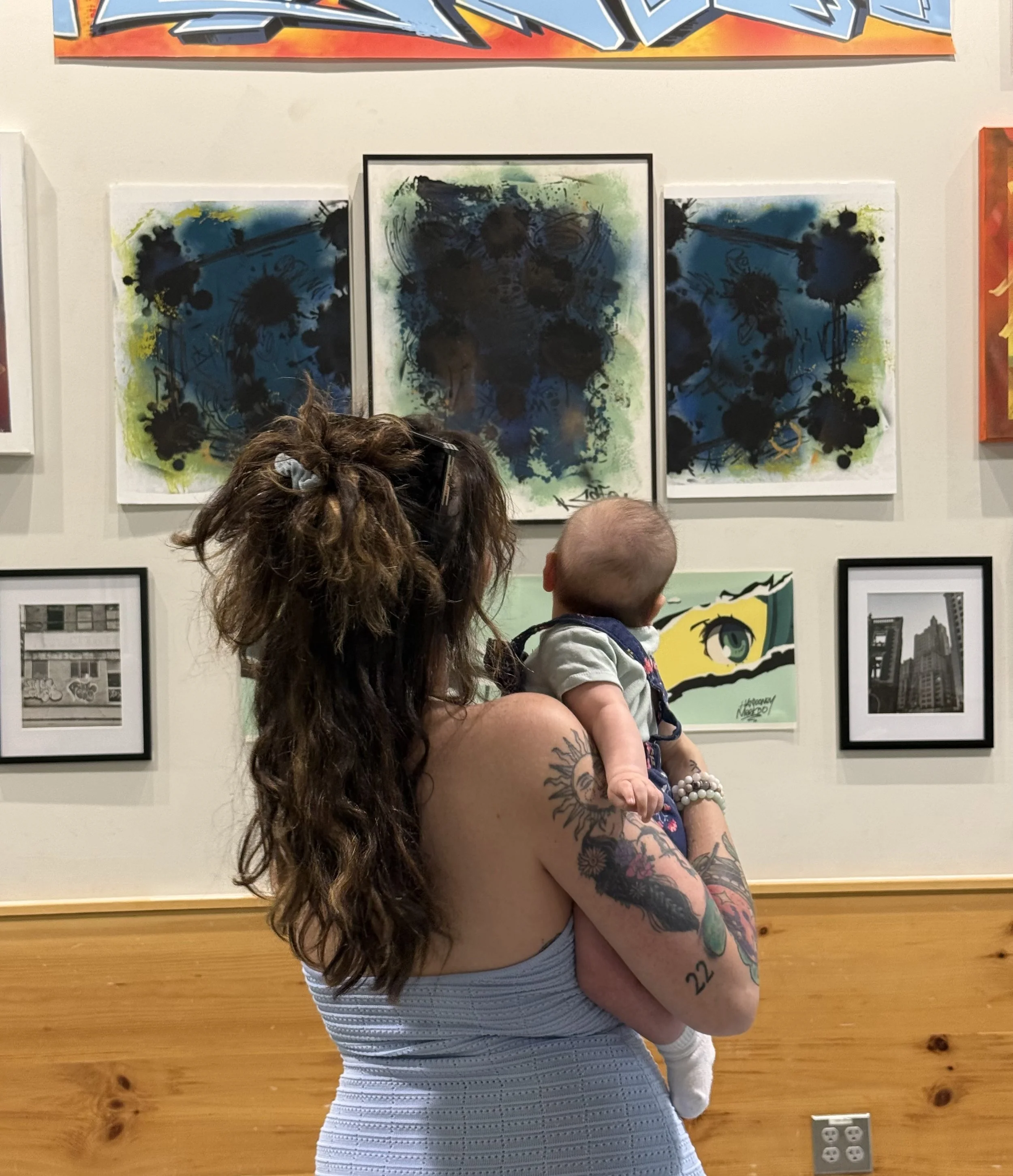 A woman with long, curly hair and tattoos on her arms is holding a baby. They are in an art gallery with colorful paintings on the wall behind them.