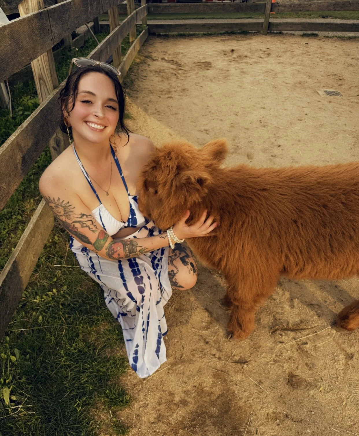 A smiling woman kneeling down and holding a brown furry calf in a fenced outdoor area.