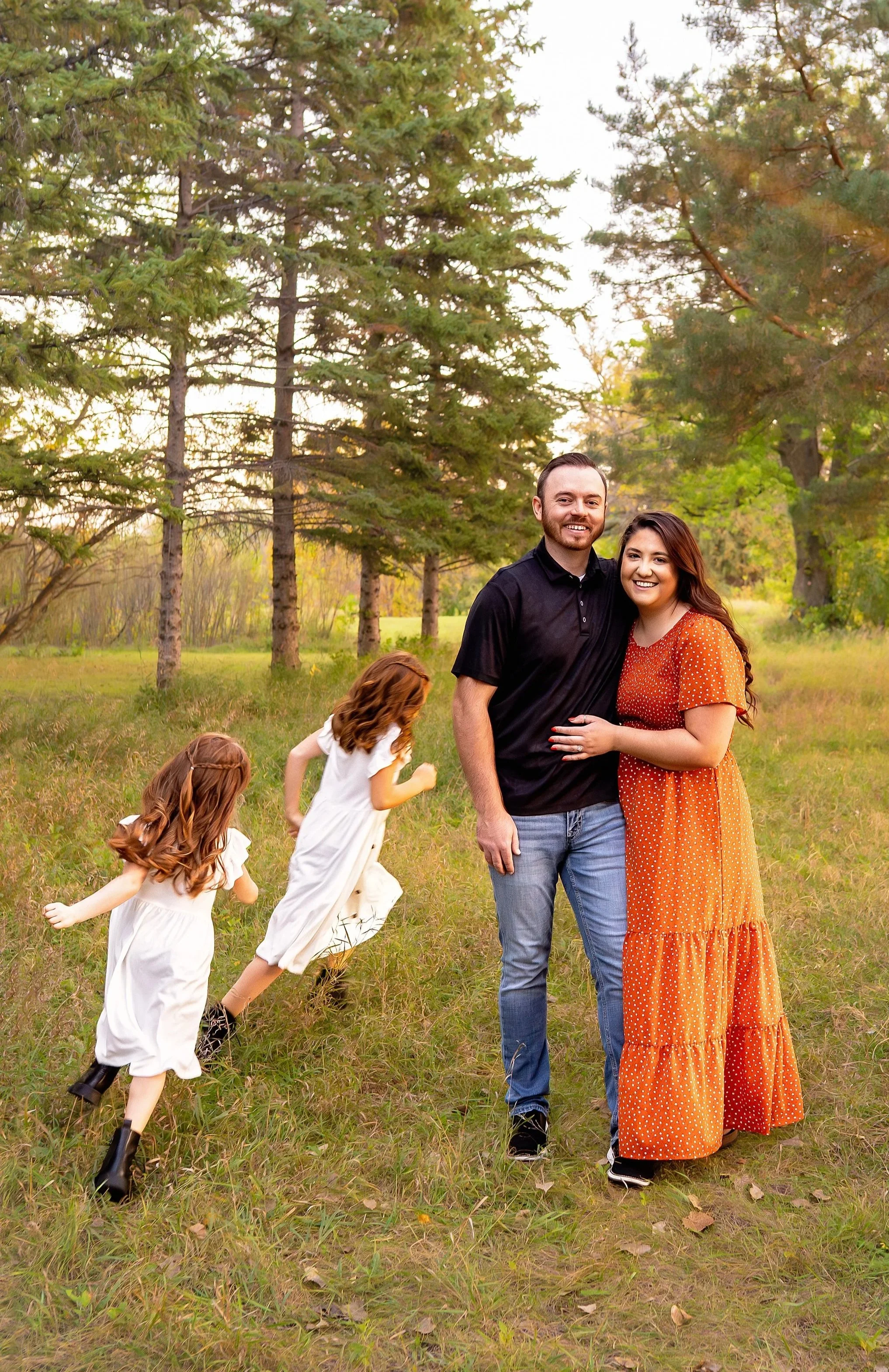 A happy couple, man with short dark hair and woman with long dark hair, standing together outdoors in a grassy area with trees in the background, smiling at the camera. Two young girls in white dresses are running and playing in front of them.