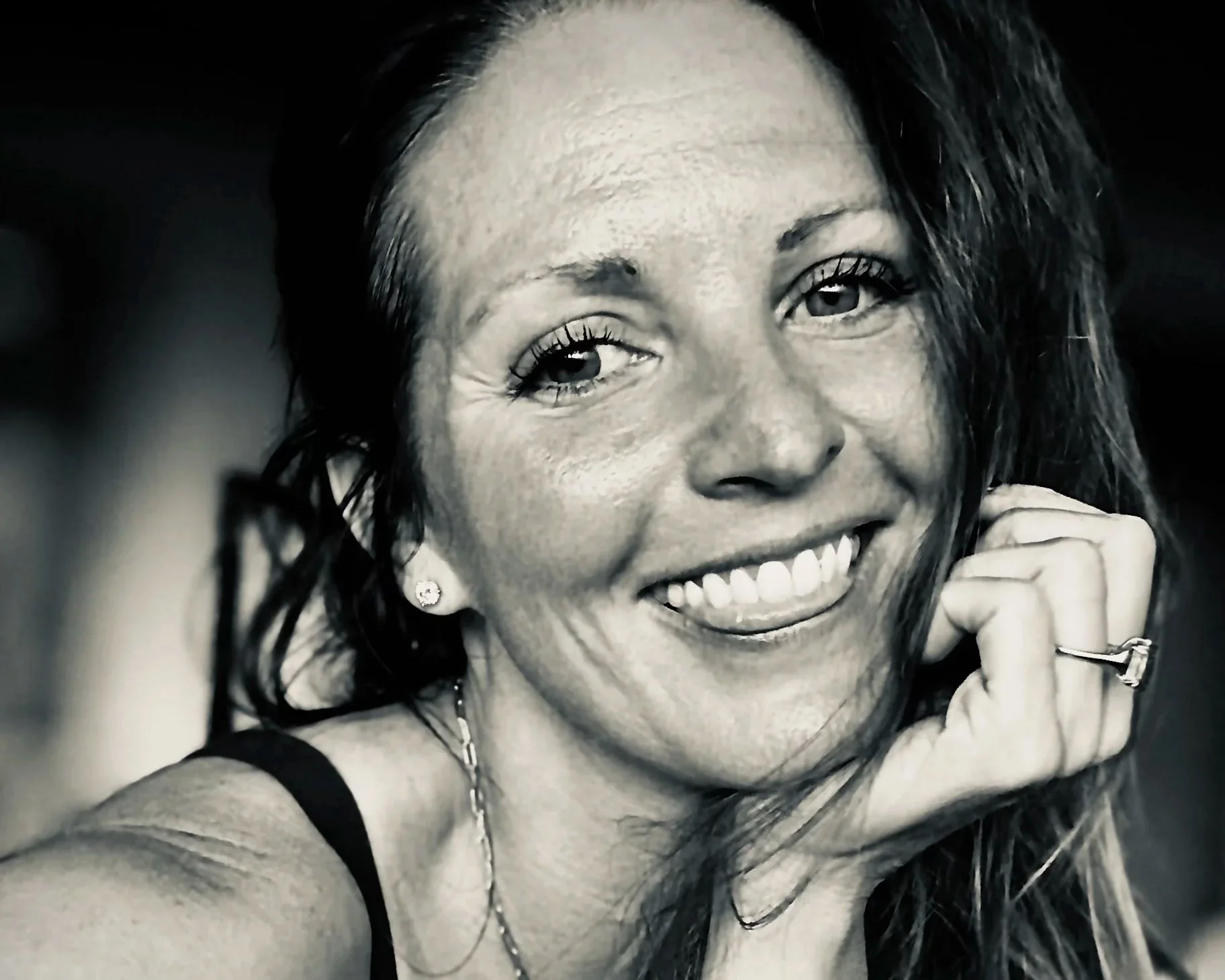 A black-and-white close-up selfie of a smiling woman with long, dark, wavy hair, wearing earrings, a ring, and a tank top, showing her teeth and dimples.