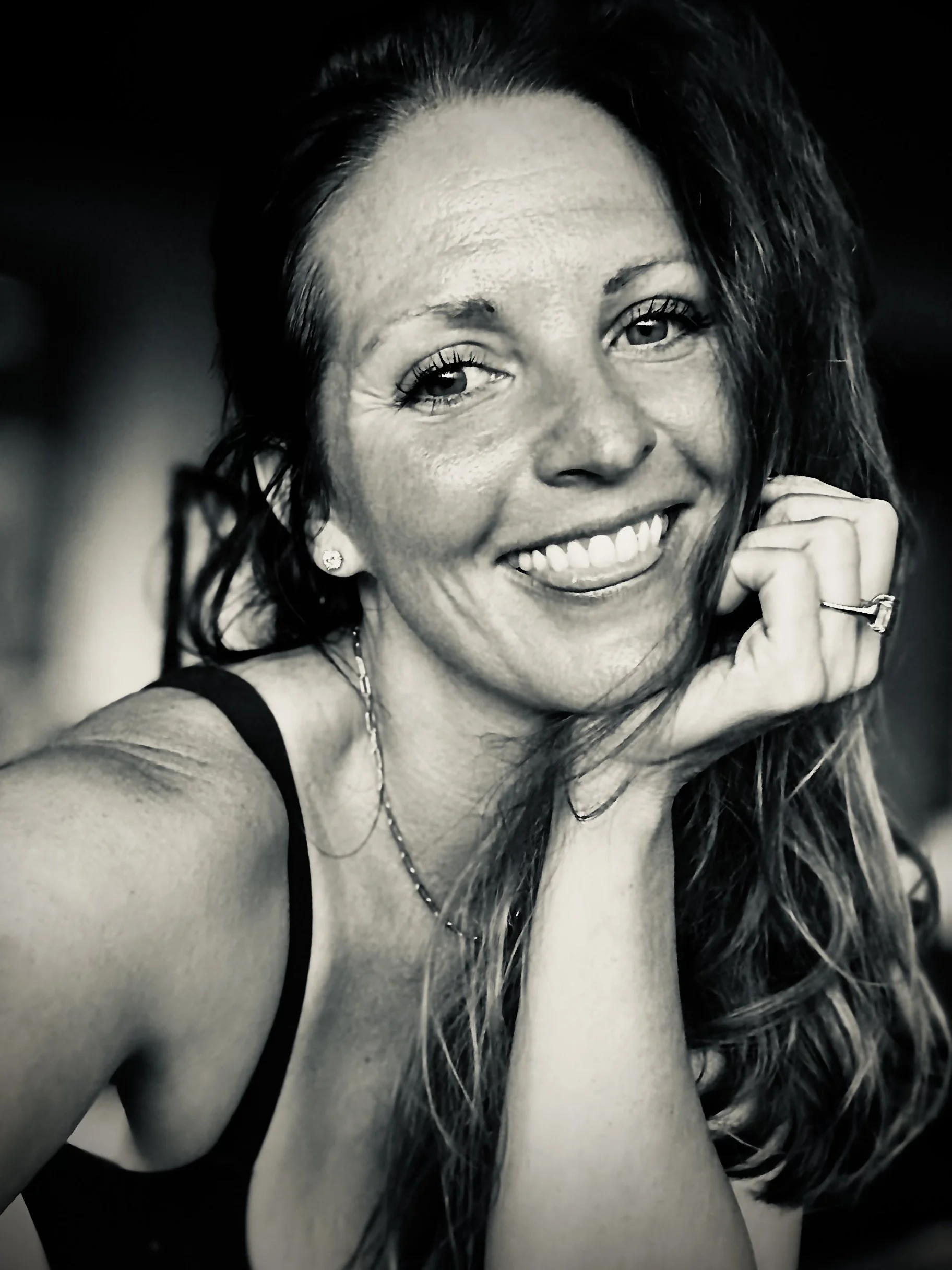 Black and white close-up of a smiling woman with wet hair, touching her face with her hand, wearing earrings and a necklace.