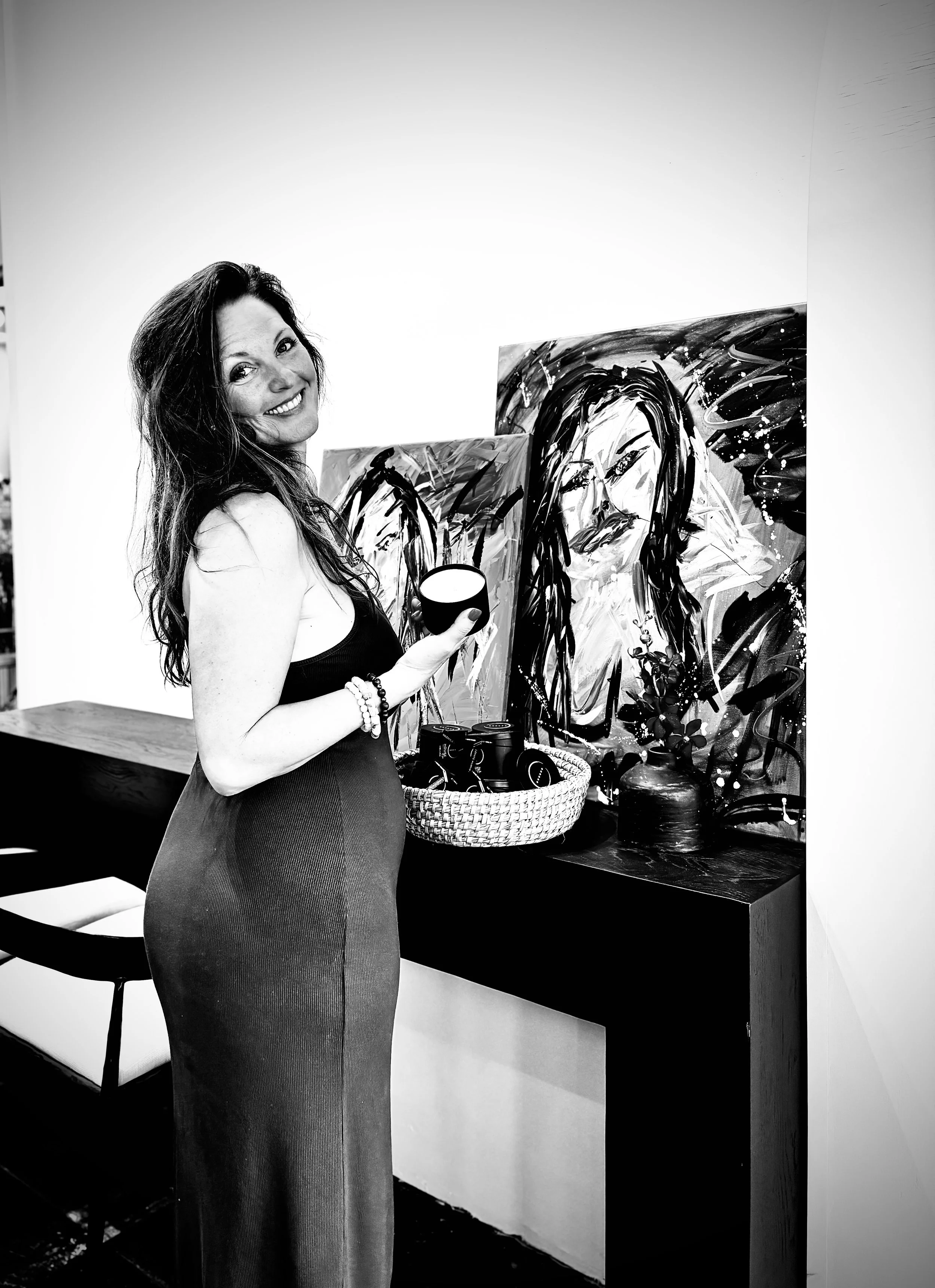 A woman with long dark hair smiling, holding a cup in her right hand, standing in front of an abstract black and white painting of a woman's face with long hair. She is wearing a sleeveless dress and bracelets, in an art gallery or exhibition space.