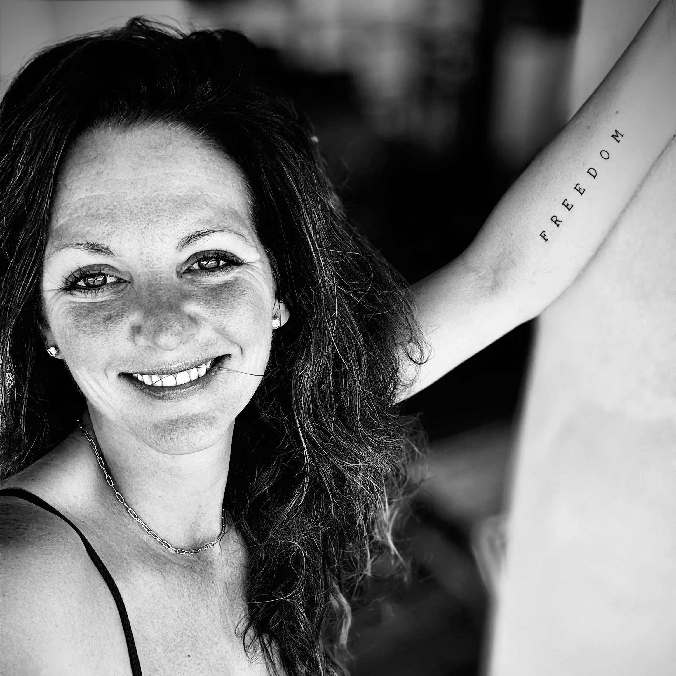 Close-up black and white photograph of a woman with dark hair smiling, wearing earrings and a chain necklace, with part of her arm visible showing a tattoo that reads 'FREEDOM'.