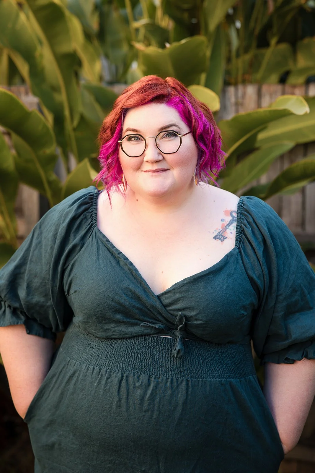 A woman with pink and brown hair, wearing glasses and a dark green dress, smiling outdoors in front of green foliage.
