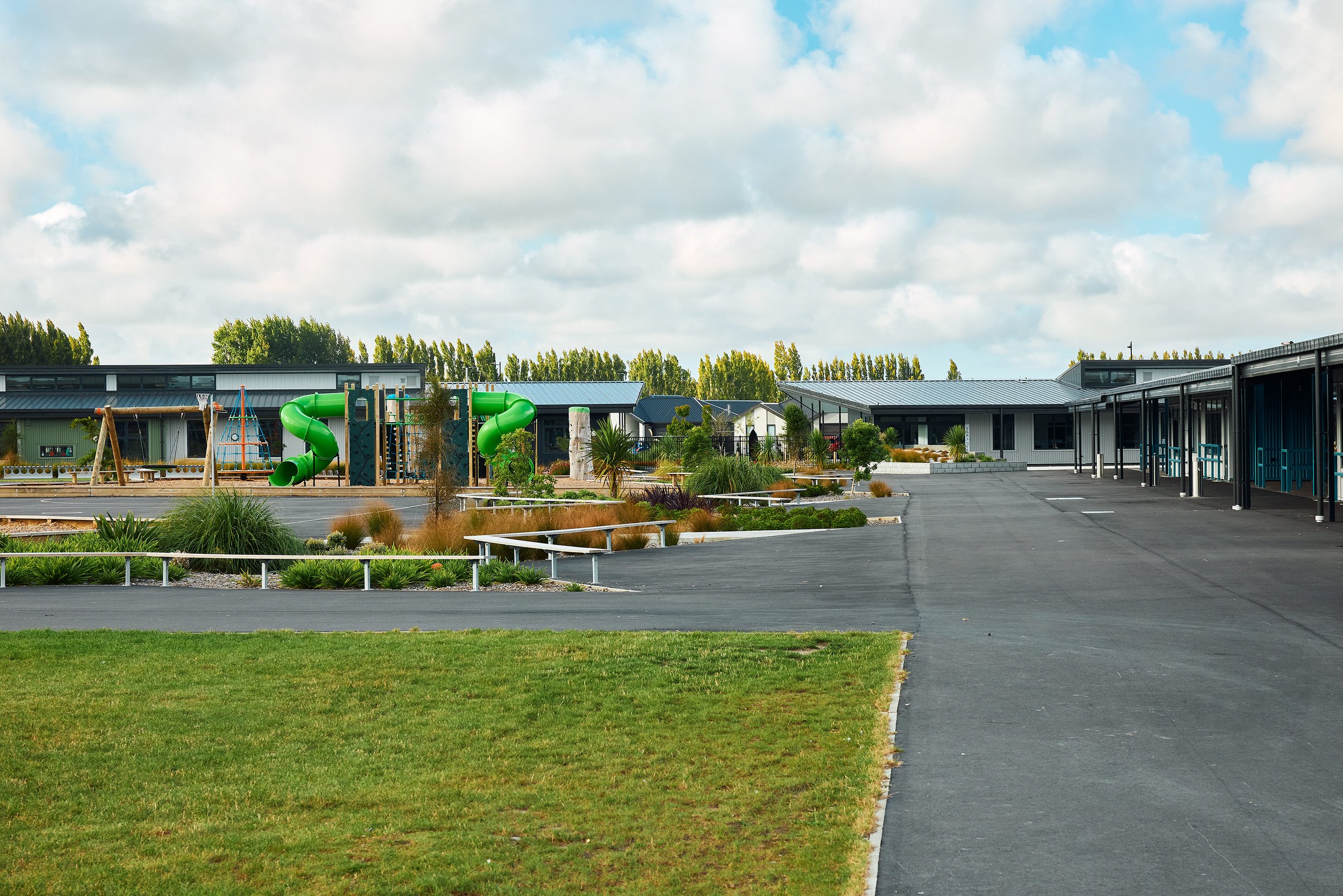 A modern school or community center with a playground, green water slide, paved parking lot, landscaping, and buildings with metal roofs under a cloudy sky.