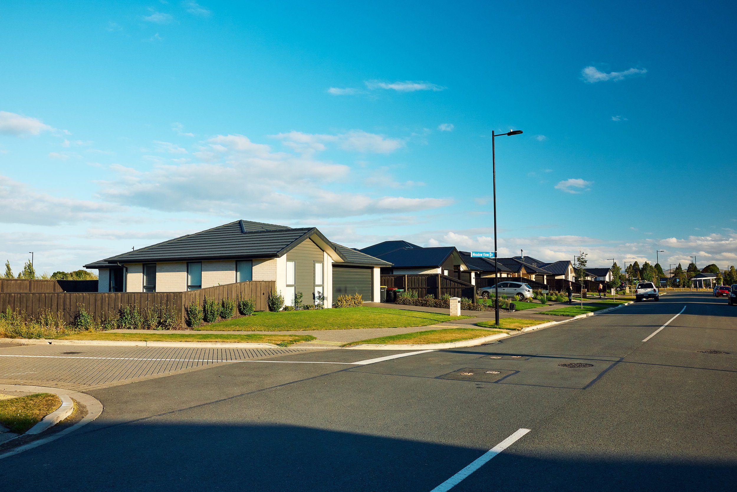 A suburban neighborhood street with houses, parked cars, and a street sign that reads 'Meadow View Dr'. The sky is blue with some clouds and there are streetlights along the road.