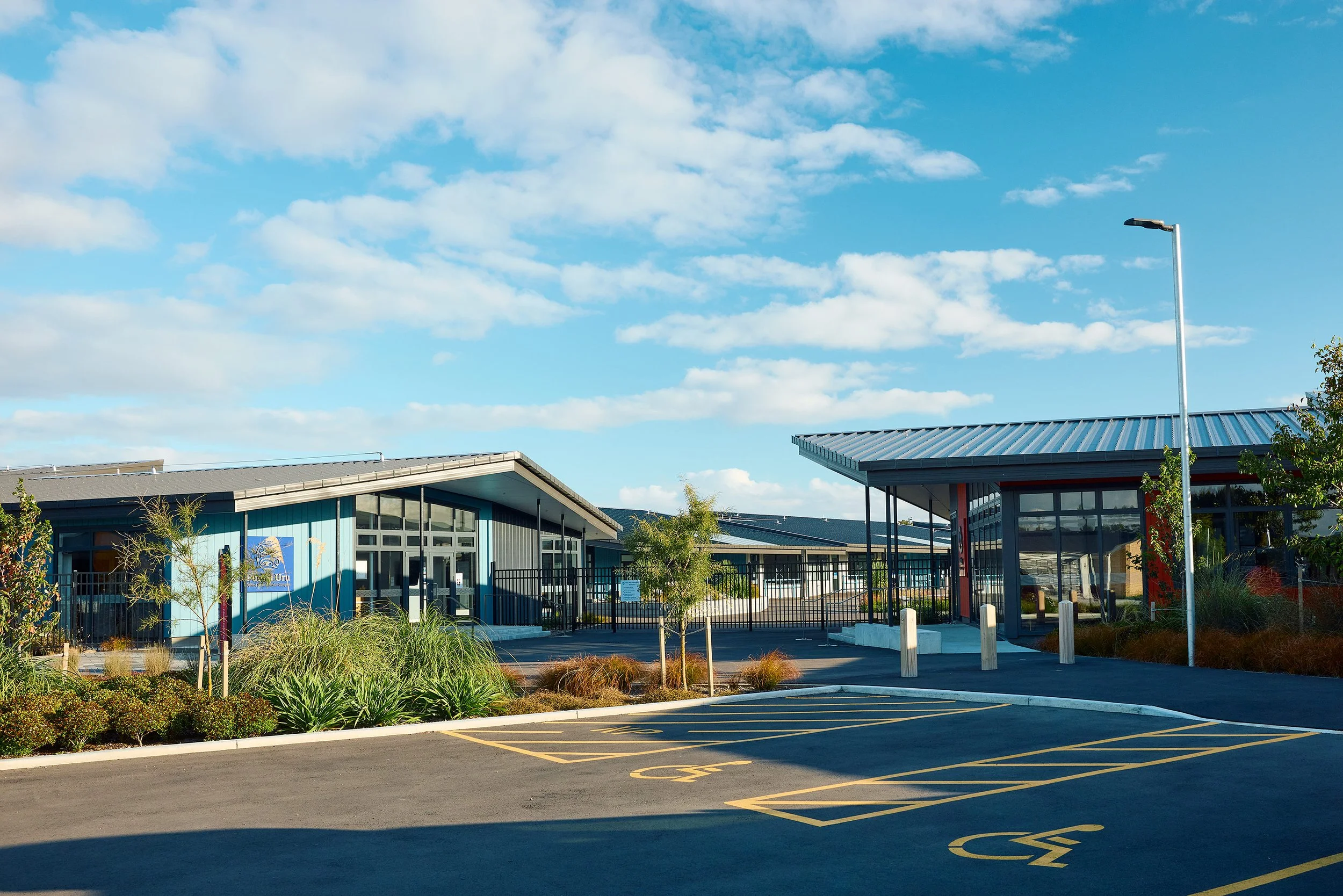 Exterior of a modern building with a blue facade, surrounded by landscaped bushes and trees. A parking lot with handicapped and regular spaces is in the foreground, and a clear blue sky with scattered clouds is overhead.
