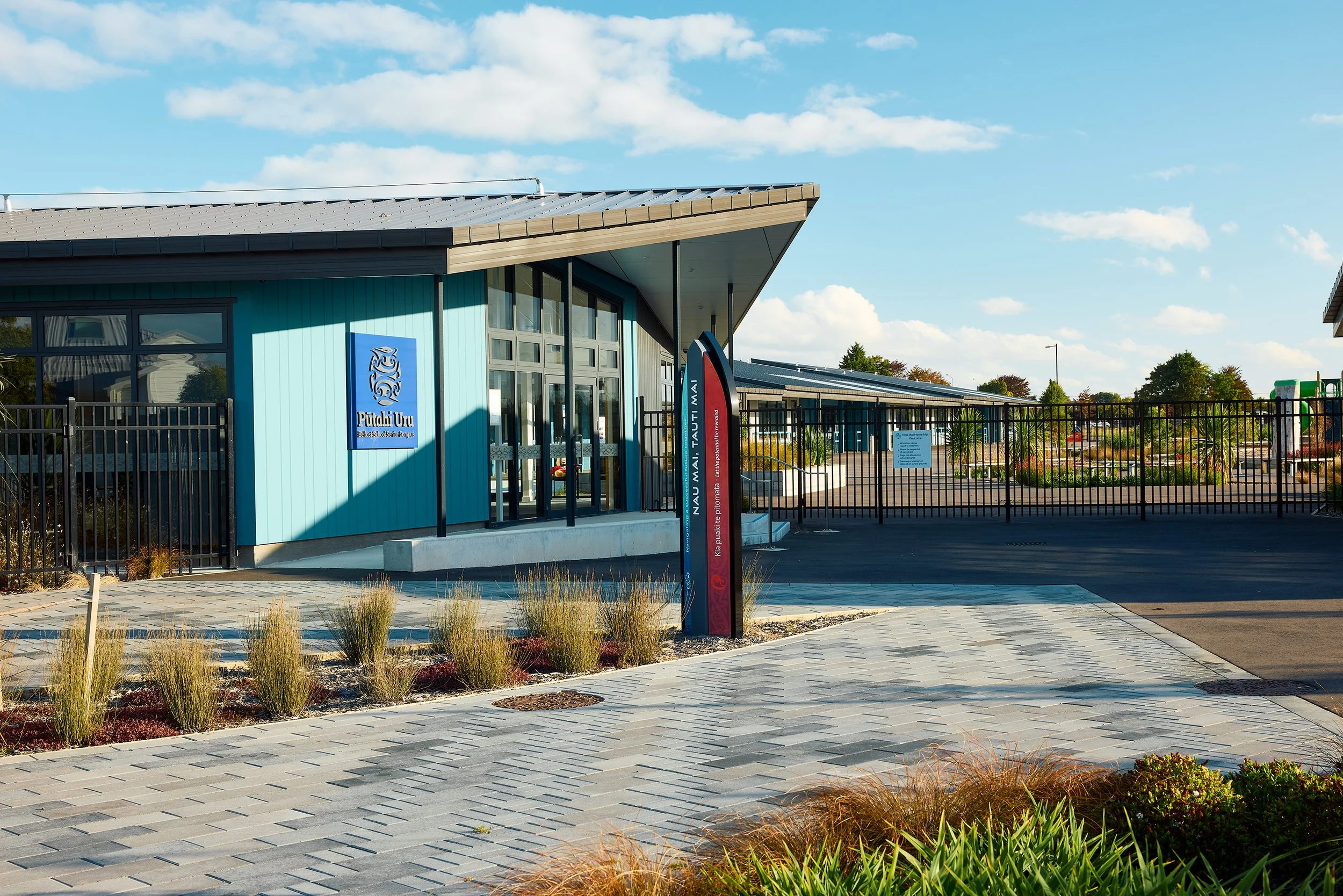 Exterior view of a modern school building with light blue siding, large windows, a metal roof, and a parking lot surrounded by a black metal fence. There is a sign that says 'Putaki Uru' and some landscaping with plants and paving stones.