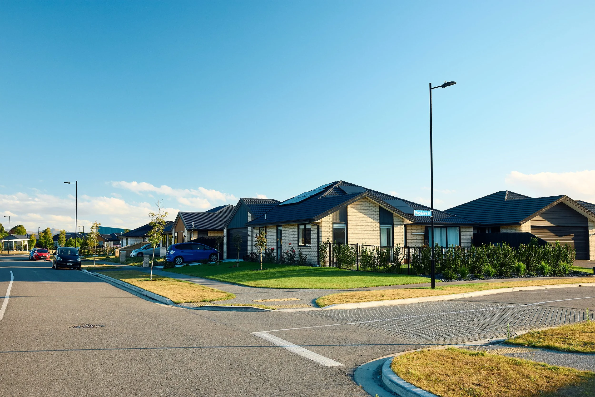 A residential neighborhood with modern houses that have dark roofs, a sidewalk, streetlights, and a parking area with several cars under a clear blue sky.