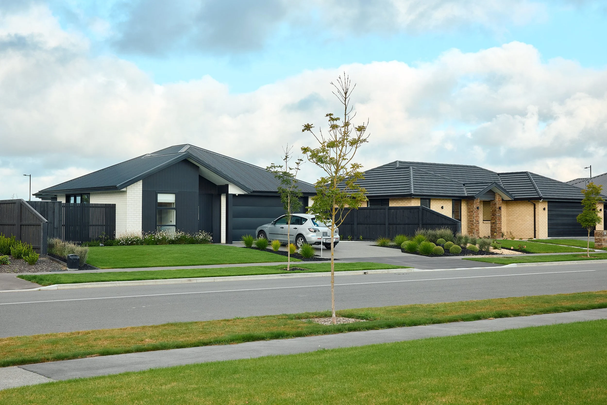 A suburban house with a dark roof and light brick exterior, surrounded by a manicured lawn with small trees and bushes, and a parked car in the driveway. A black fence runs along the yard, with a street in the foreground and a cloudy sky above.