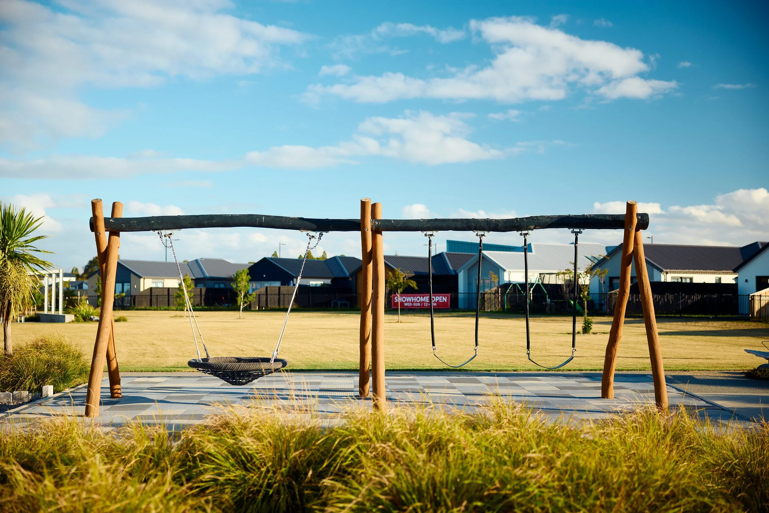 Playground swings in a park with houses and a blue sky in the background.