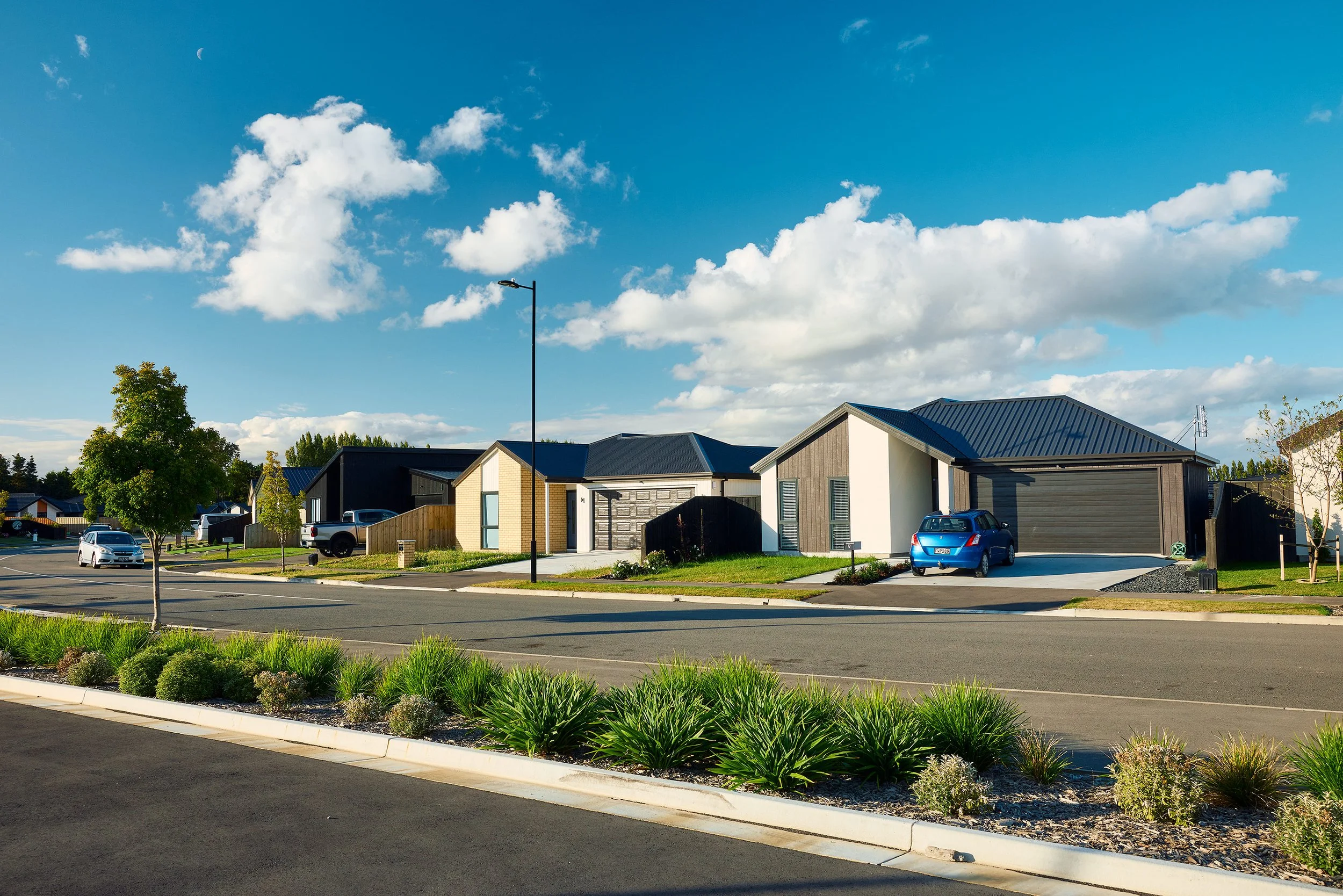 A suburban neighborhood street with modern houses, parked cars, green lawns, and a blue sky with clouds and a visible moon.