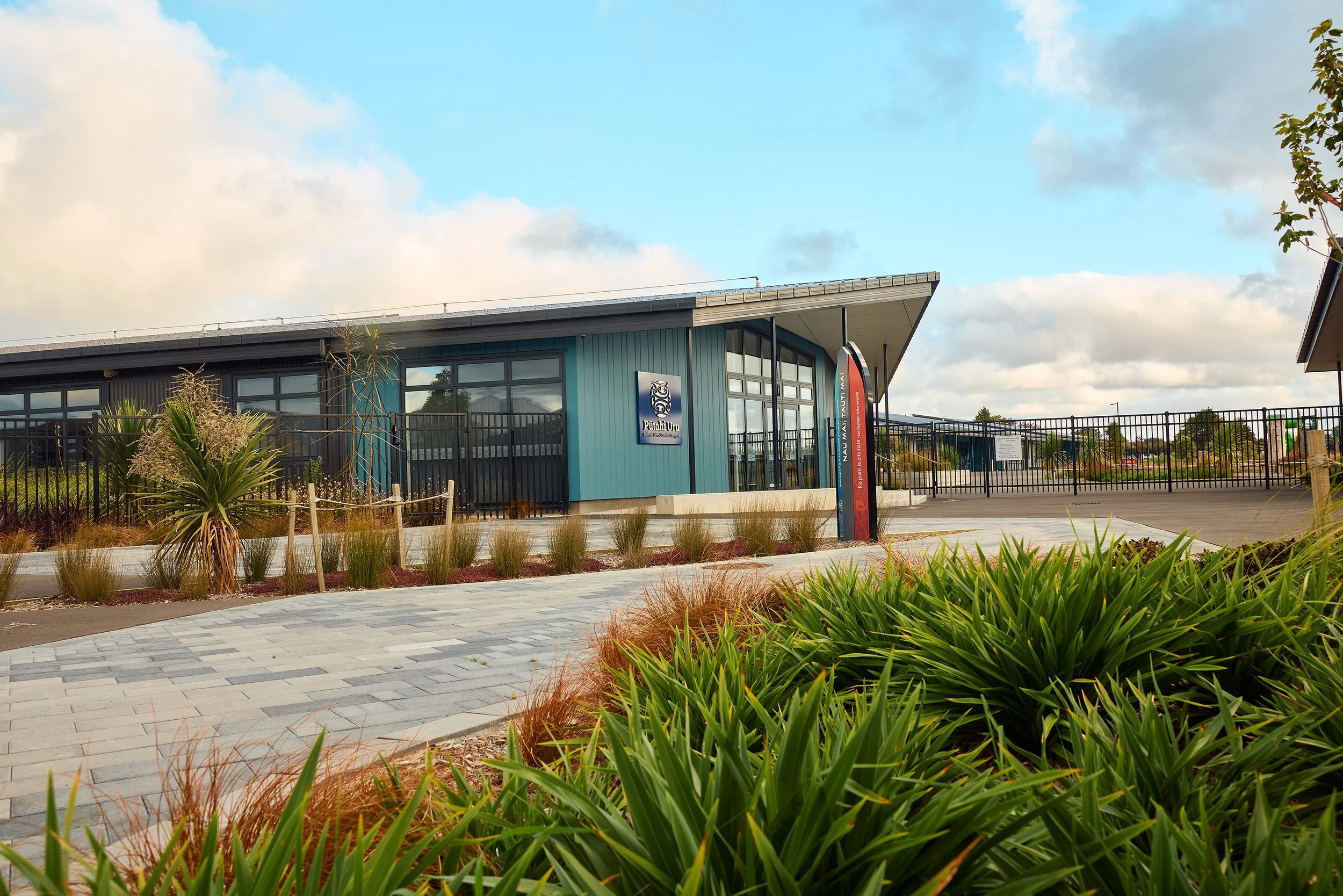 Modern building with blue exterior, large windows, and signs, surrounded by landscaped plants and paved walkway under a partly cloudy sky.