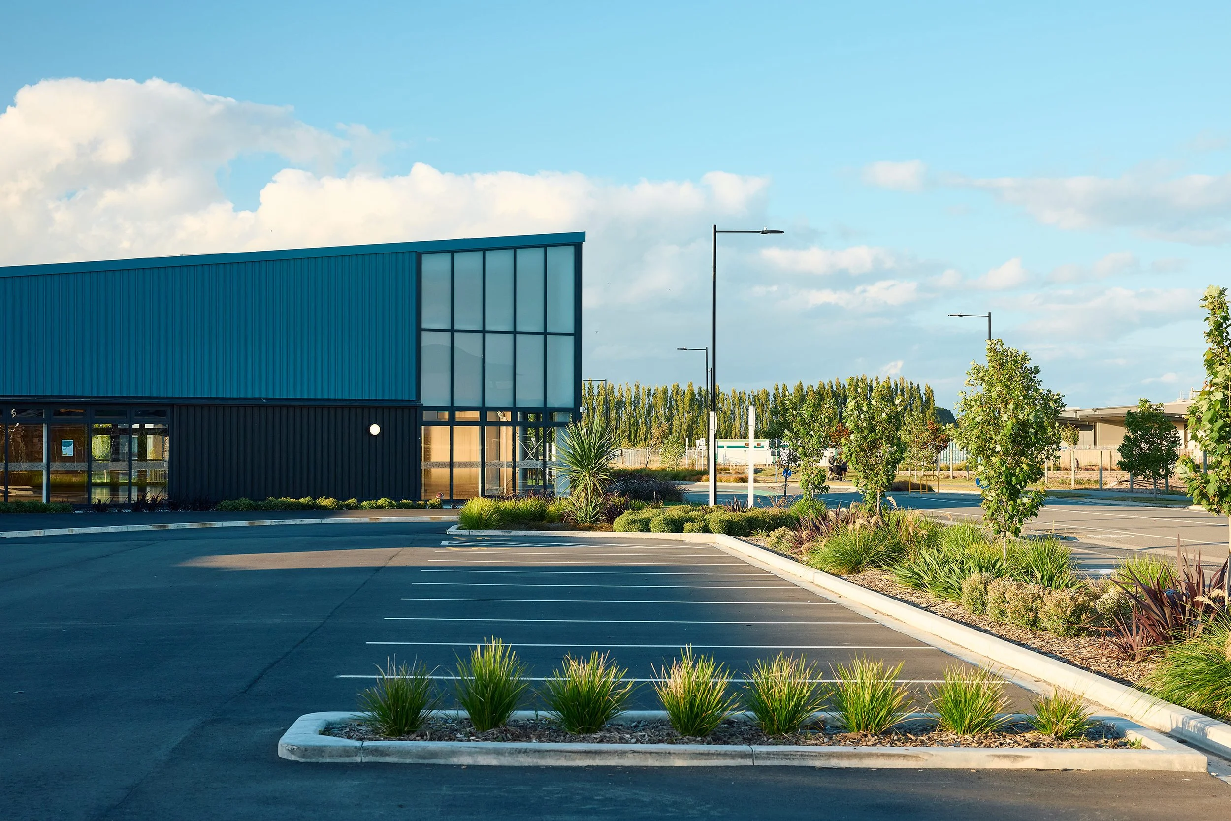 An empty parking lot in front of a modern blue building with large windows, surrounded by landscaped greenery and trees, under a blue sky with scattered clouds.