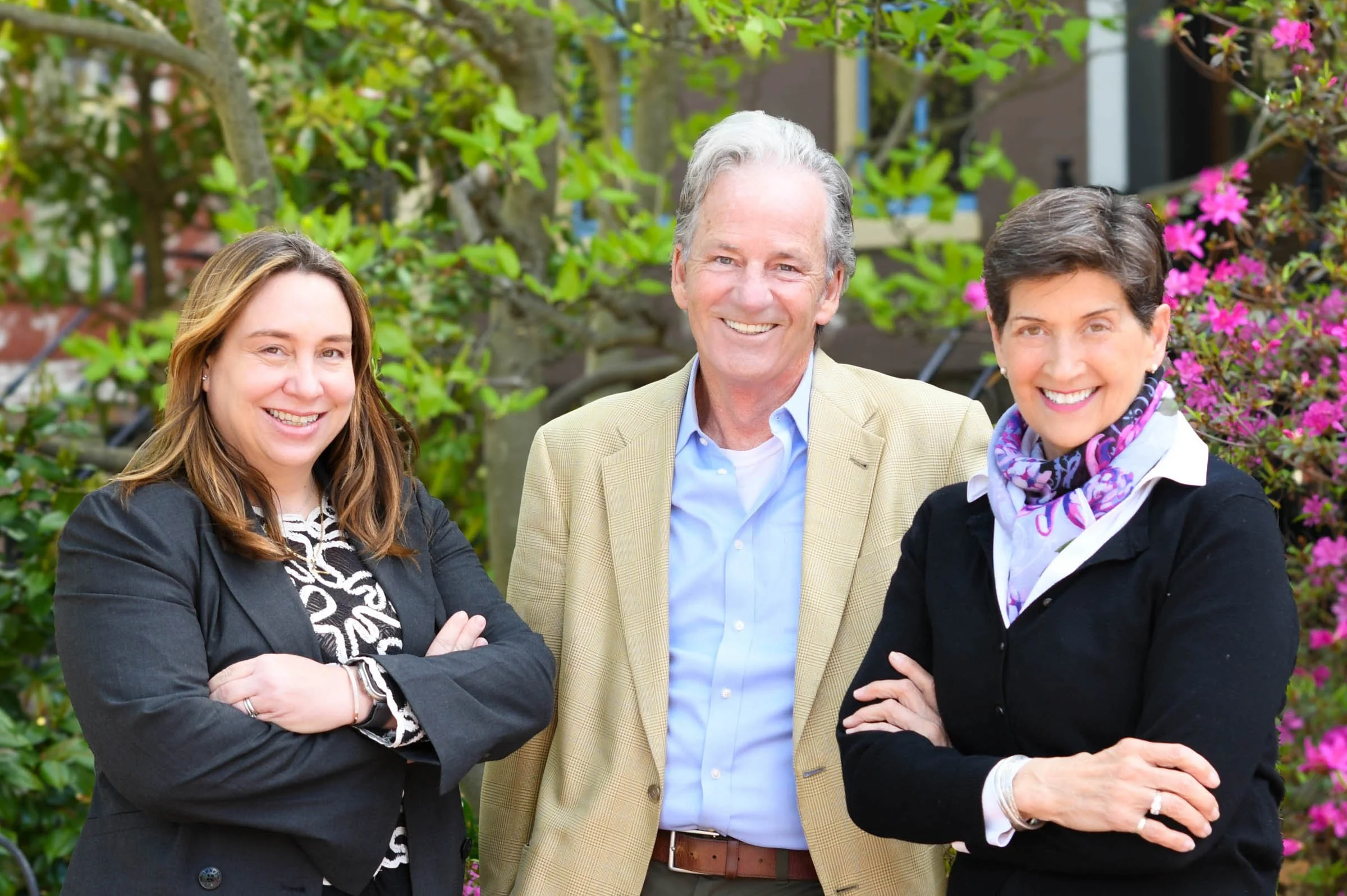 Three people standing outdoors with greenery and pink flowering bushes in the background. They are smiling, with two women on either side and a man in the middle.