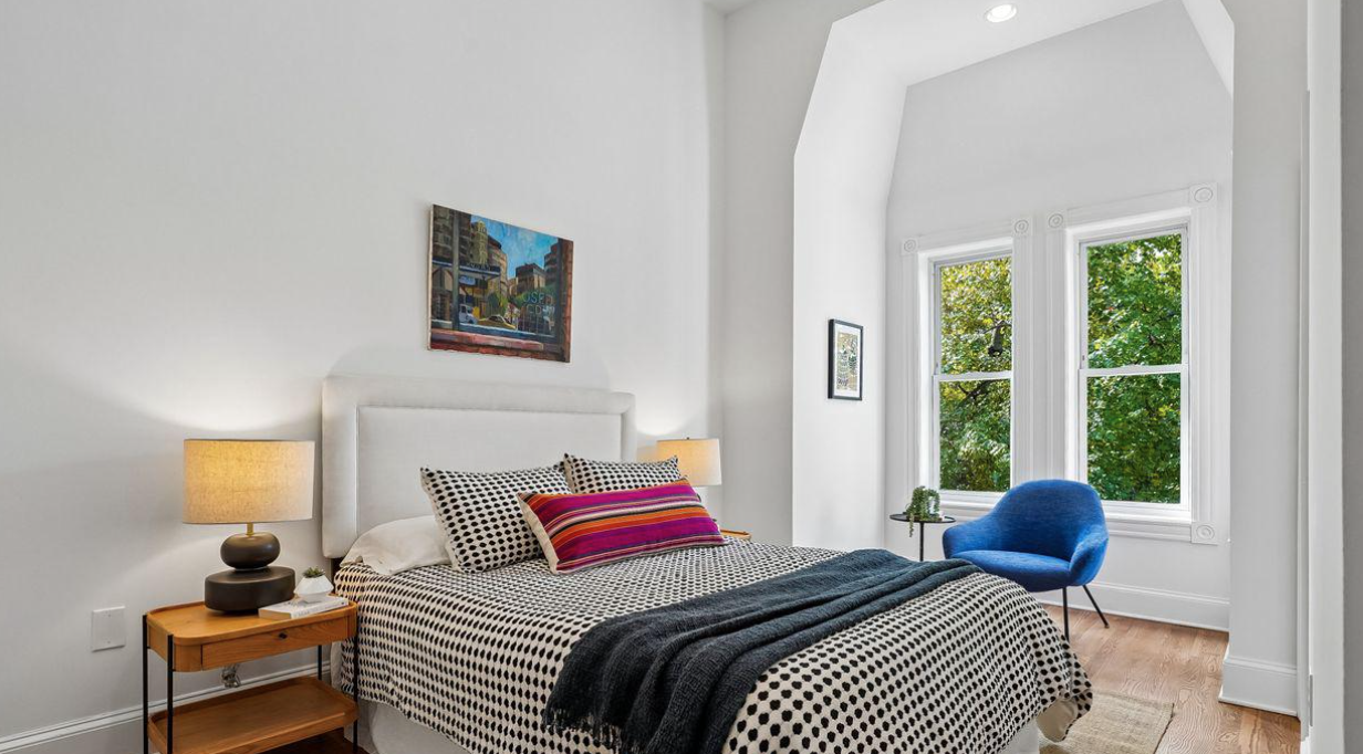 A bright bedroom with white walls and a large window showing green trees outside. The bed has a white headboard, black and white polka dot bedding, and colorful pillows. There are two lamps on a small wooden nightstand and a blue armchair near the window.
